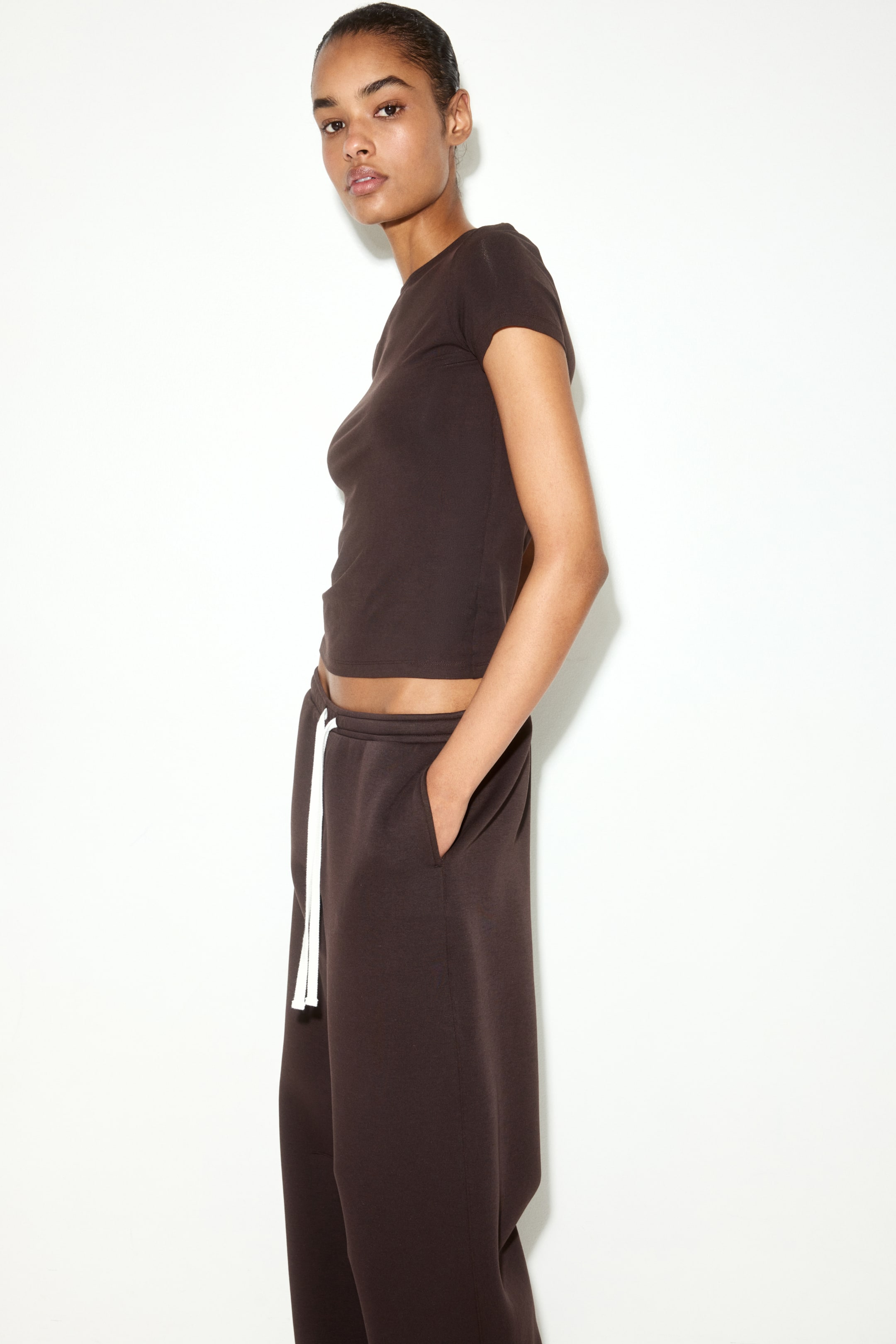 View larger image: A dark brown fitted t-shirt featuring a crew neck, short sleeves, and a straight-cut, cropped hem is worn by the model, who pairs it with matching brown sweatpants.