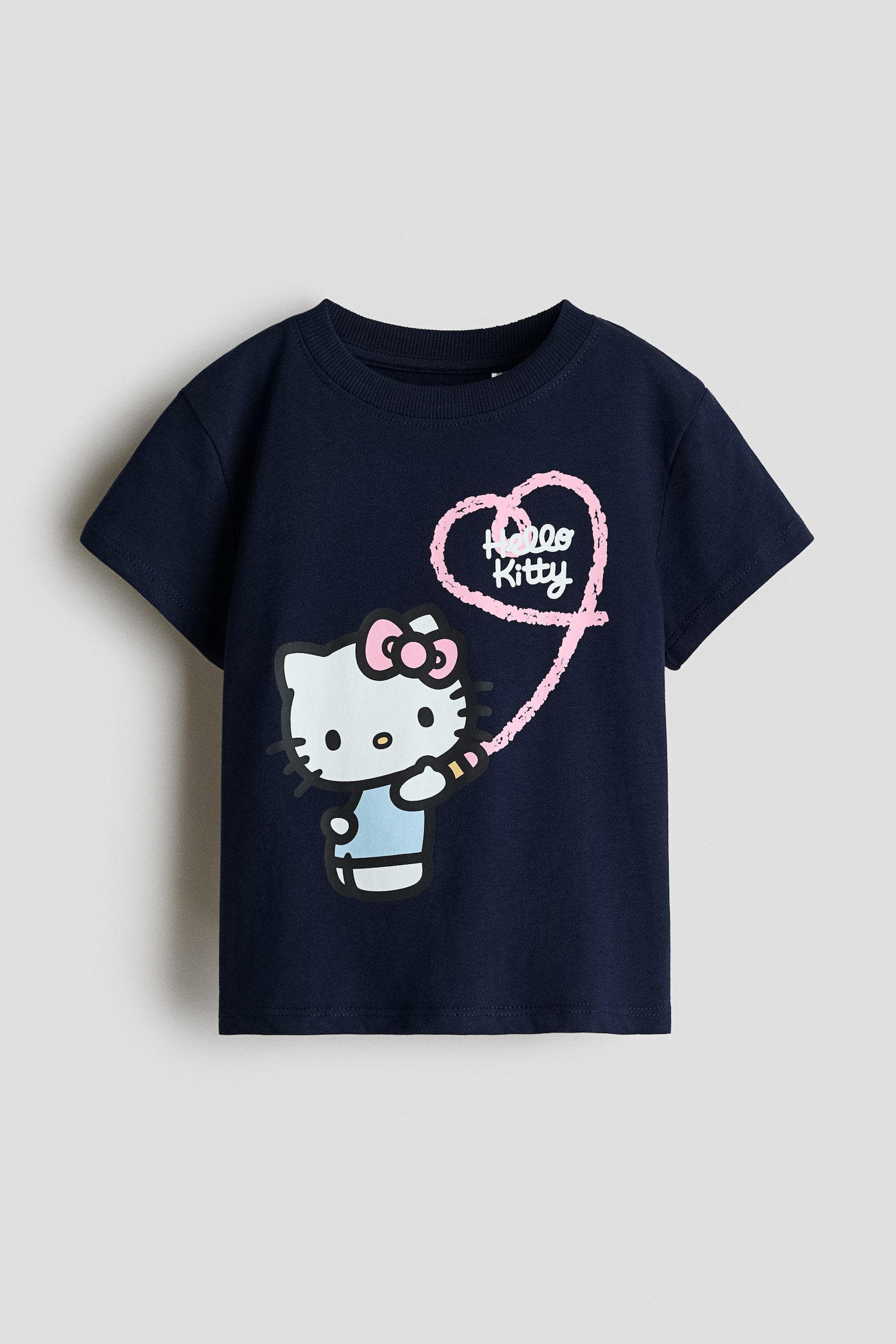A navy blue cotton jersey T-shirt is shown from the front, featuring a round, rib-trimmed neckline and short sleeves with dropped shoulders. The front displays a graphic print of Hello Kitty holding a pink pencil and drawing a pink heart containing the words "Hello Kitty". - 1