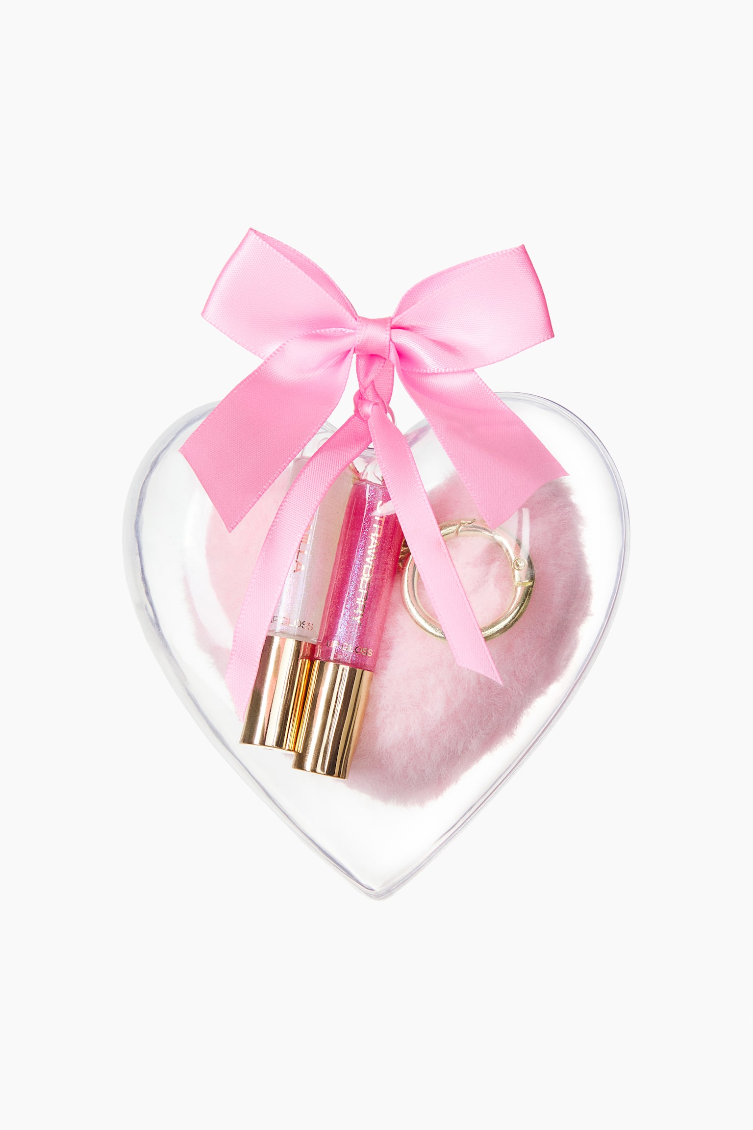 Two shimmering lip glosses with gold-coloured caps are resting inside a clear, heart-shaped plastic bauble alongside a fluffy pink pom-pom and a gold-tone metal clasp. A pink satin ribbon is tied in a bow at the top loop of the container.