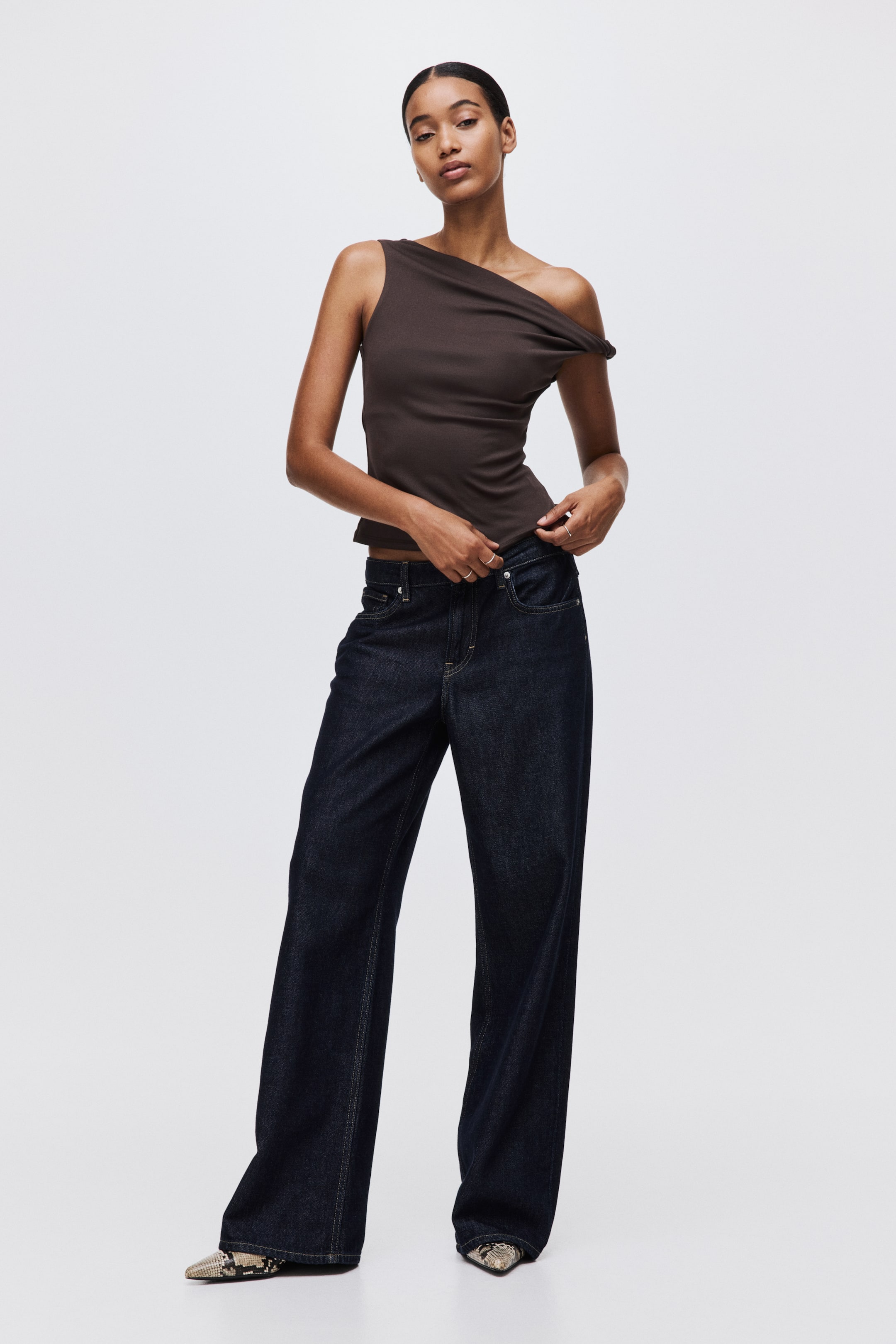 View larger image: A dark brown asymmetrical one-shoulder top is being presented from a front angle, featuring a fitted silhouette with a strap on the right shoulder and a draped design over the left. It is being paired with dark wash, high-waisted wide-leg jeans, showcasing a relaxed fit through the leg.