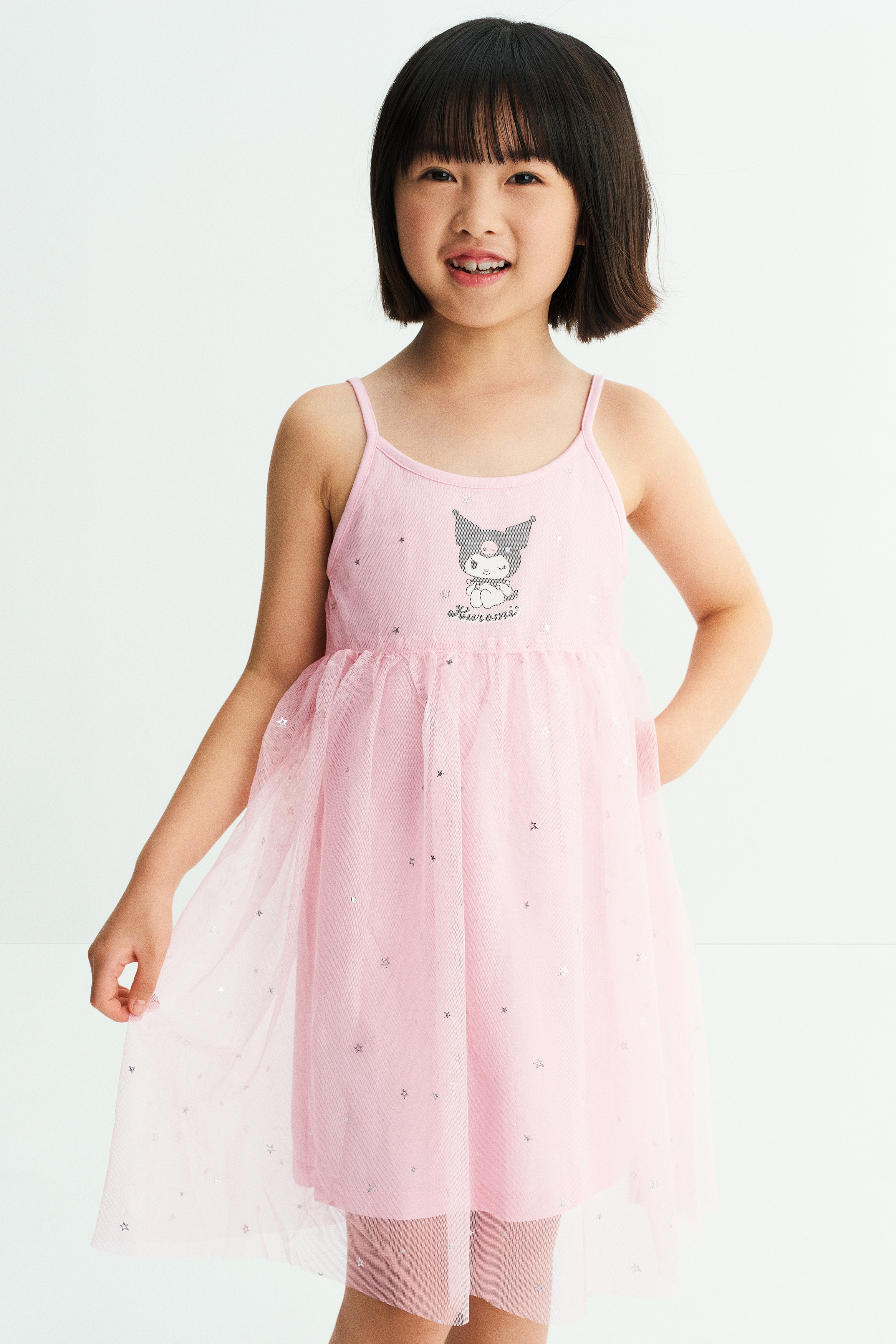 A light pink jersey dress is shown from the front, featuring narrow shoulder straps, a round neckline, and a grey Kuromi character print on the chest. The dress features a sheer, gathered tulle skirt overlay decorated with small silver stars over a solid pink jersey base.