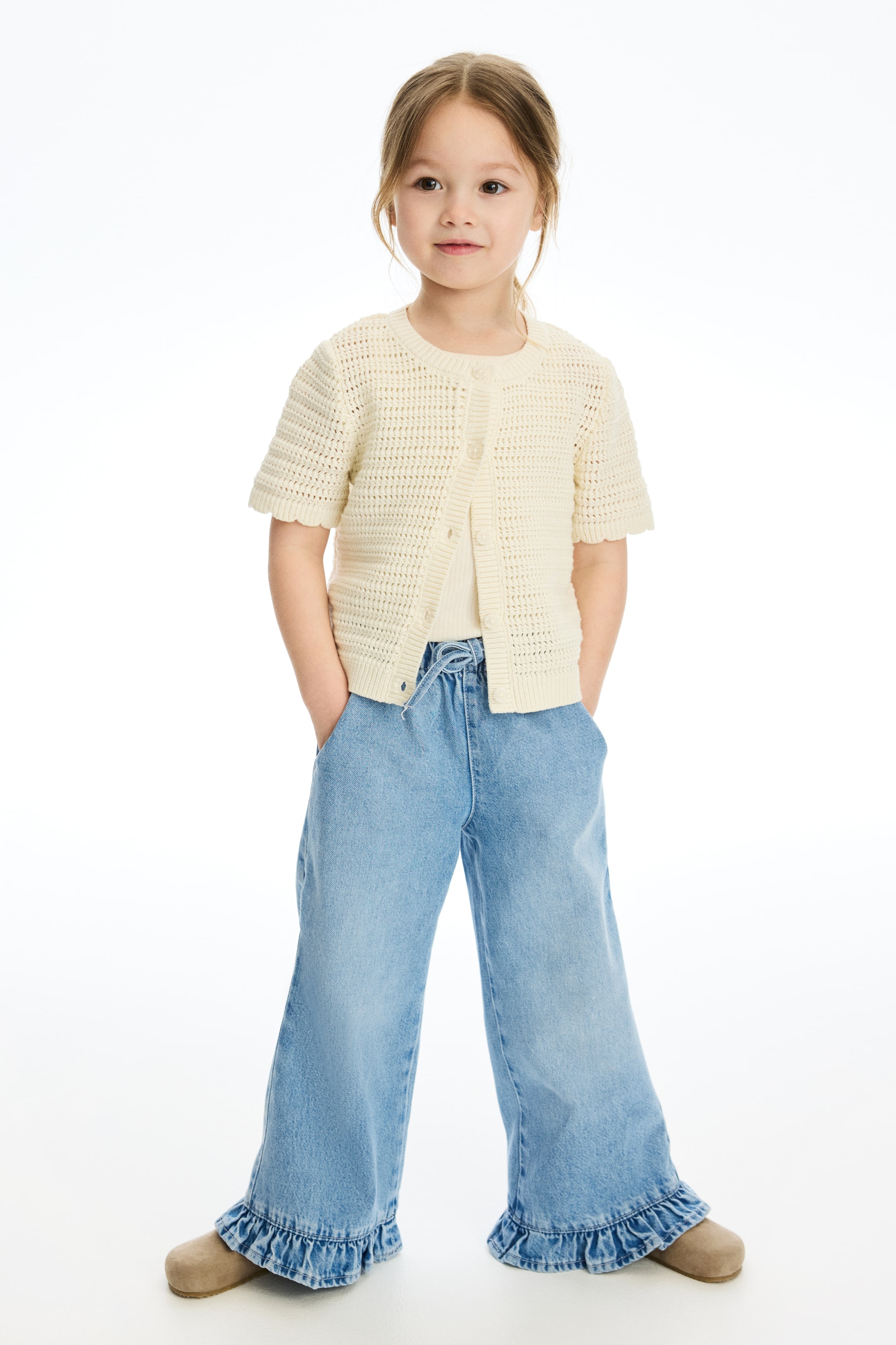 View larger image: The cream short-sleeved cardigan is crafted with an open-knit design, a round neckline, and a button-front closure. It features scalloped edges on the sleeves and hem, and is styled over a light-coloured top.
