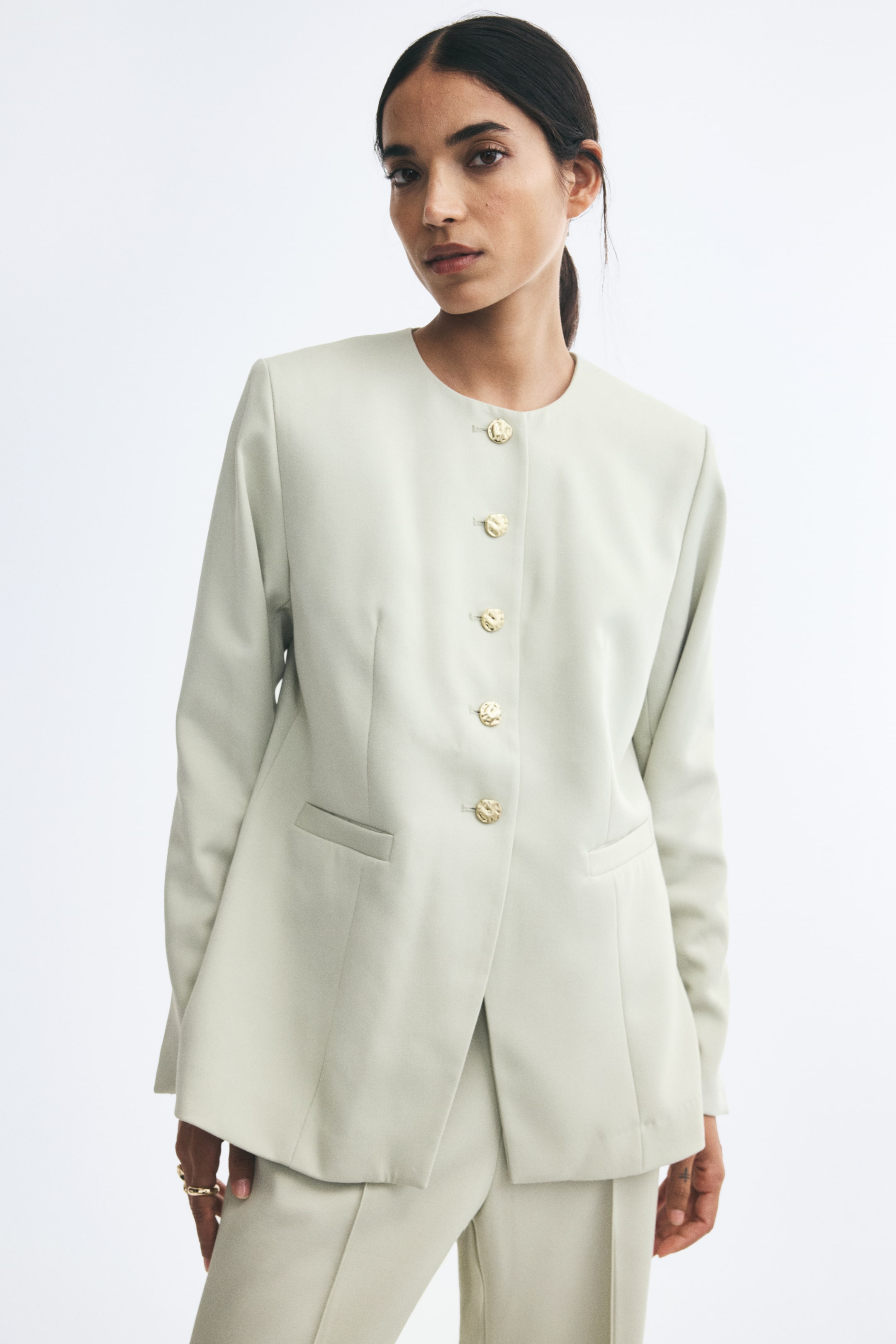 View larger image: The model is wearing a long, single-breasted twill blazer in a light sage green, featuring a collarless round neckline, structured shoulders, and irregular-shaped gold metal buttons along the front. The blazer is styled with matching trousers.
