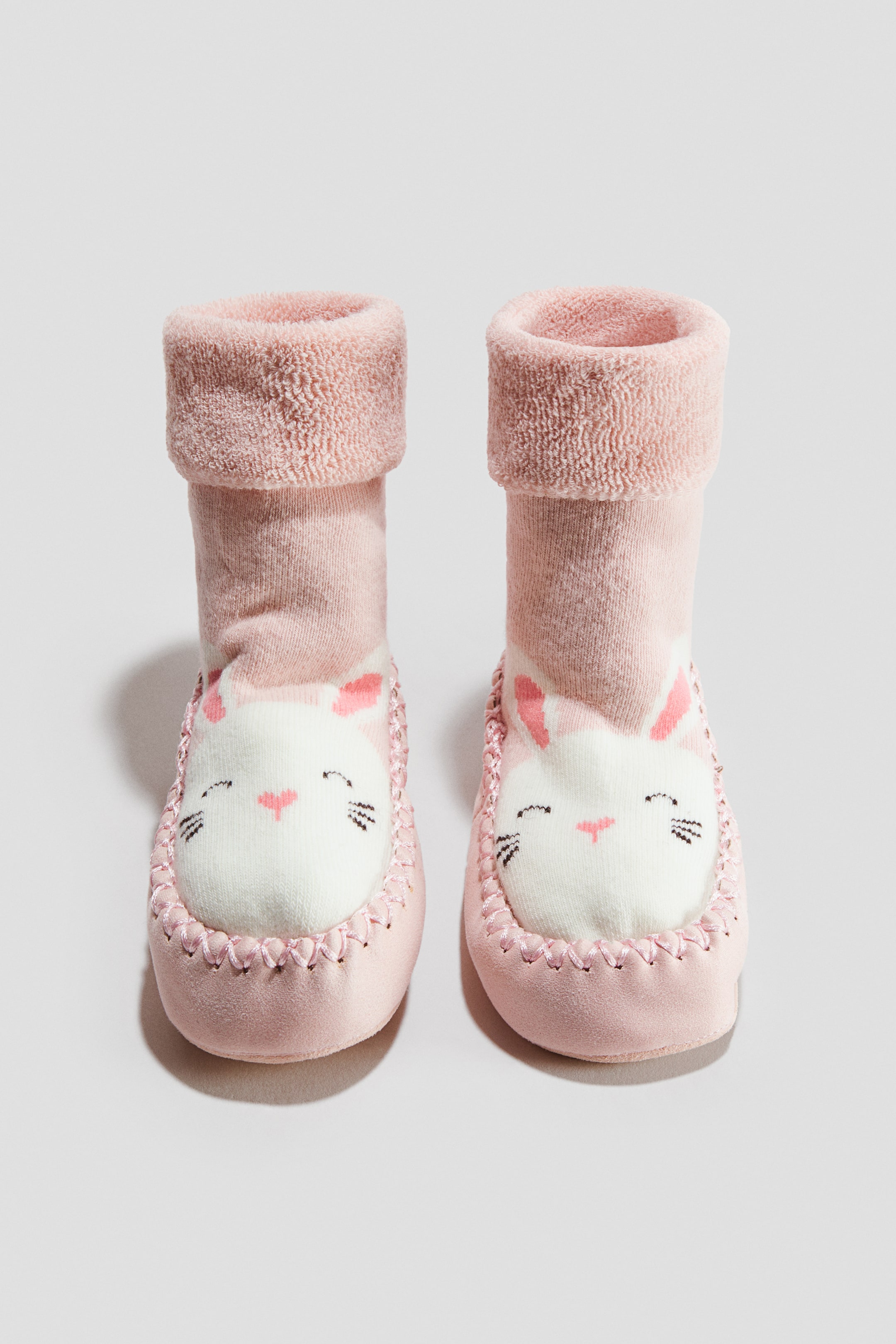 Voir une image plus grande: Une paire de petites bottines pour bébé roses est debout, bien droites, orientées vers l’avant, avec une tige de type chaussette munie d’un revers roulé moelleux et une base de style mocassin. L’avant de chaque bottine présente un empiècement blanc brodé d’un visage de chat et souligné par des coutures décoratives roses.