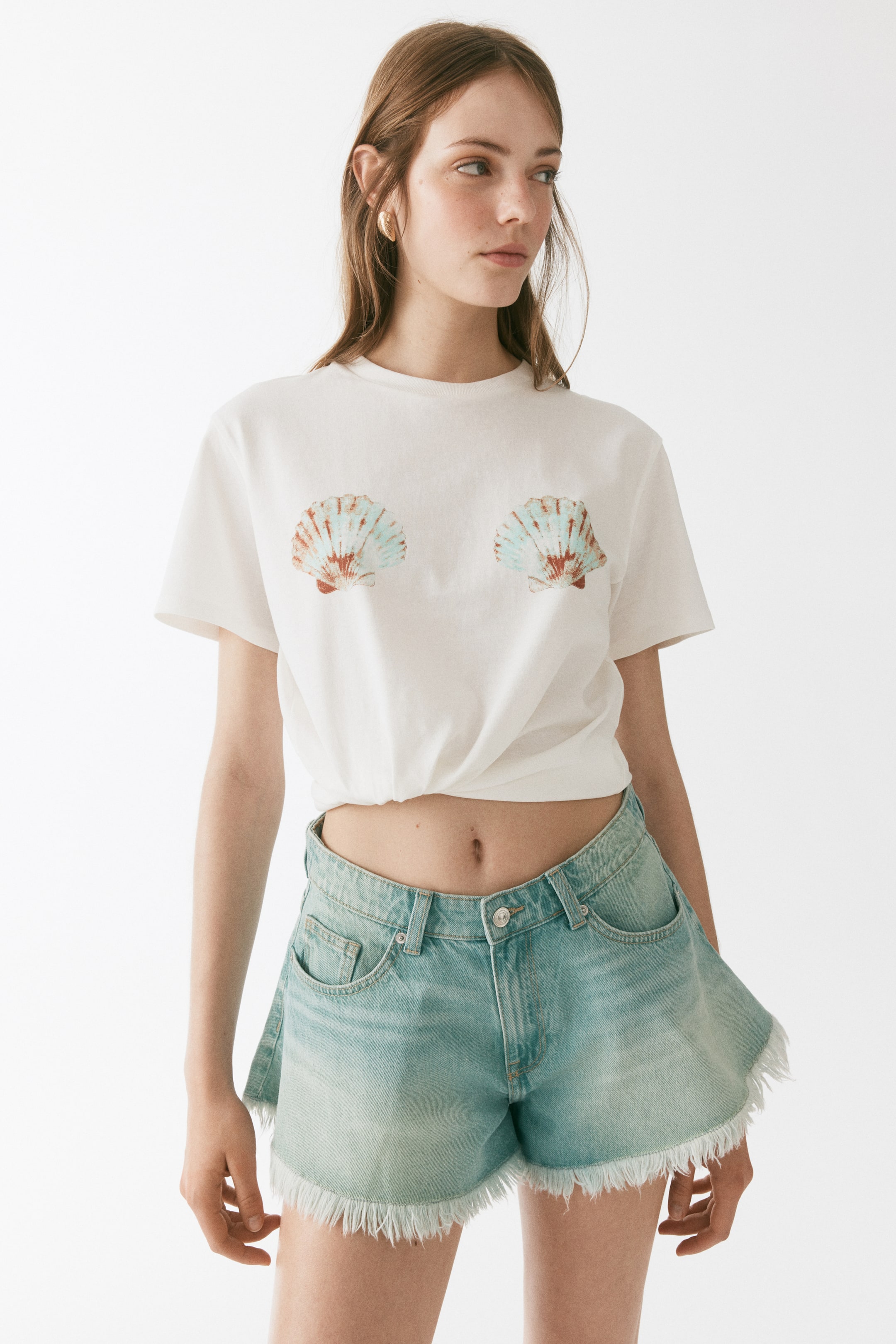 Visualizza l'immagine ingrandita: Una t-shirt bianca a girocollo è indossata, caratterizzata da due grafiche di conchiglie a ventaglio in azzurro chiaro e arancione sul davanti e l'orlo annodato. Vengono presentati anche shorts in denim lavaggio chiaro con orlo sfrangiato e tasche frontali visibili.