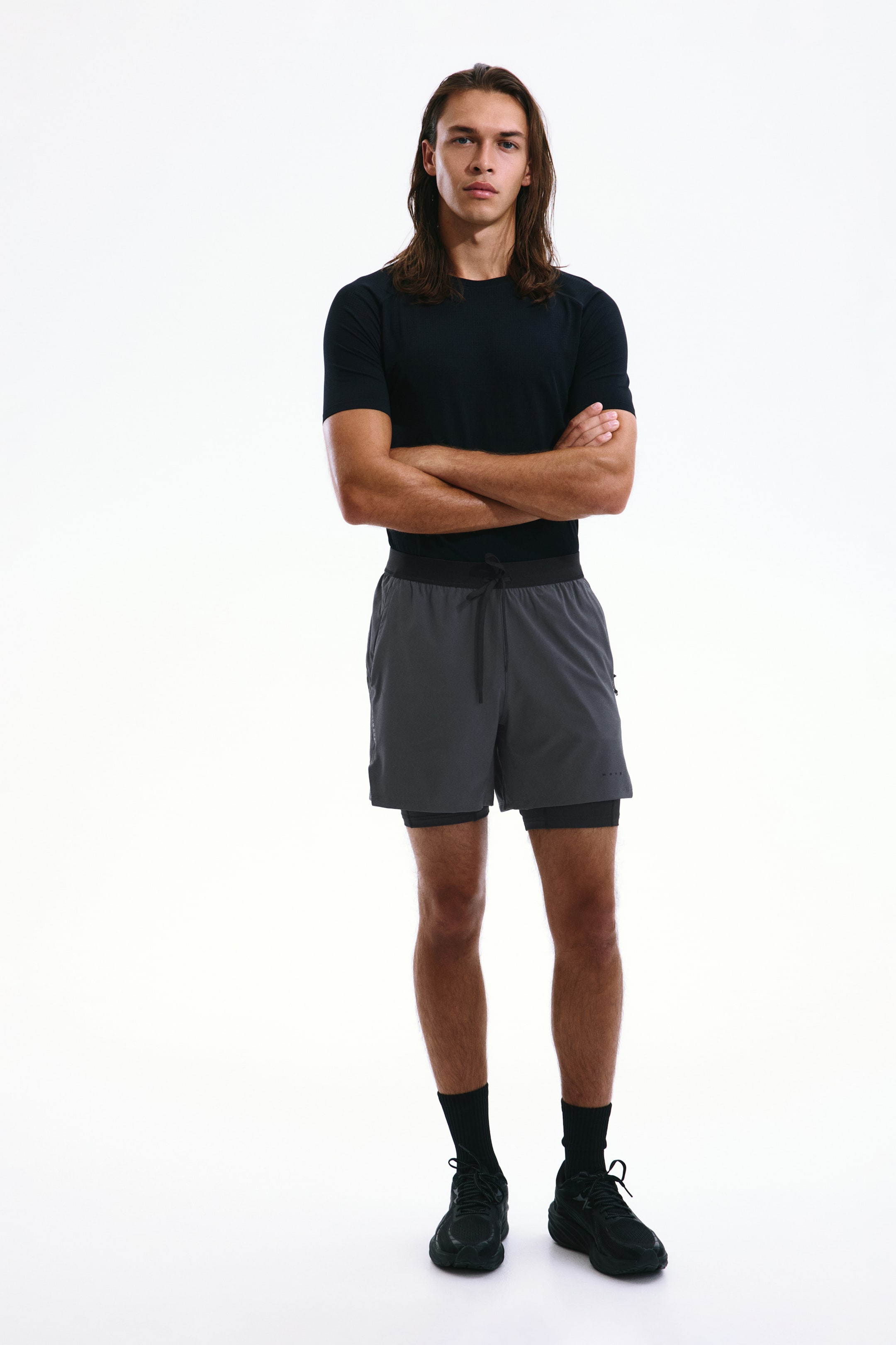 Visualizza immagine più grande: Shorts sportivi a doppio strato con DryMove indossati dalla modella, con uno strato esterno grigio scuro sopra uno strato interno aderente nero e un cordino nero in vita. La modella indossa anche un top nero a maniche corte e scarpe sportive nere.