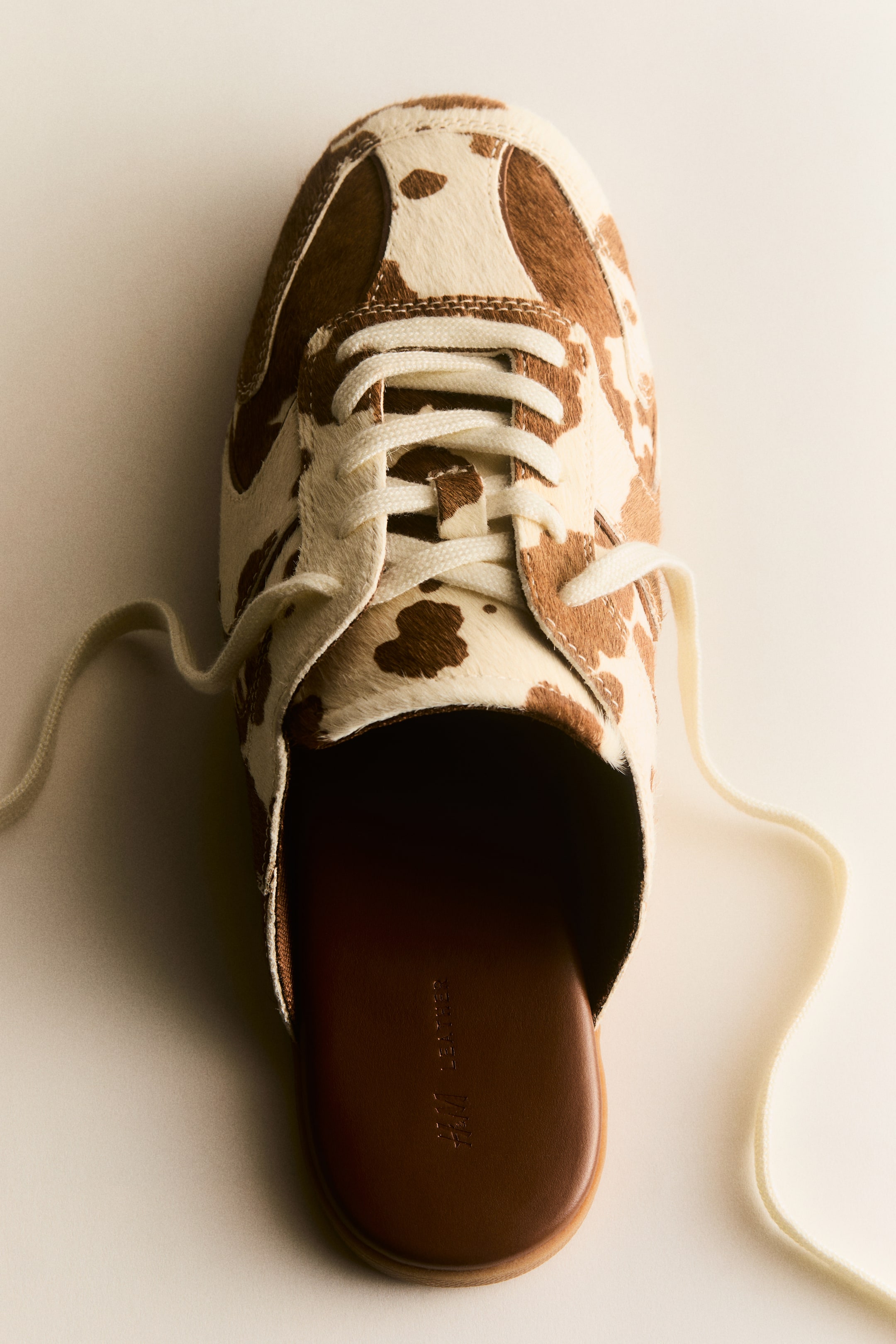 Ver imagen más grande: Un mule de estilo deportivo se presenta desde un ángulo cenital, con una parte superior de piel estampada de vaca en tonos marrón y crema, con un acabado texturizado y afelpado. Se aprecia un cordón blanco en la parte delantera sobre una lengüeta acolchada, y en el interior se muestra una plantilla de cuero marrón oscuro, grabada con 'H&M LEATHER'.