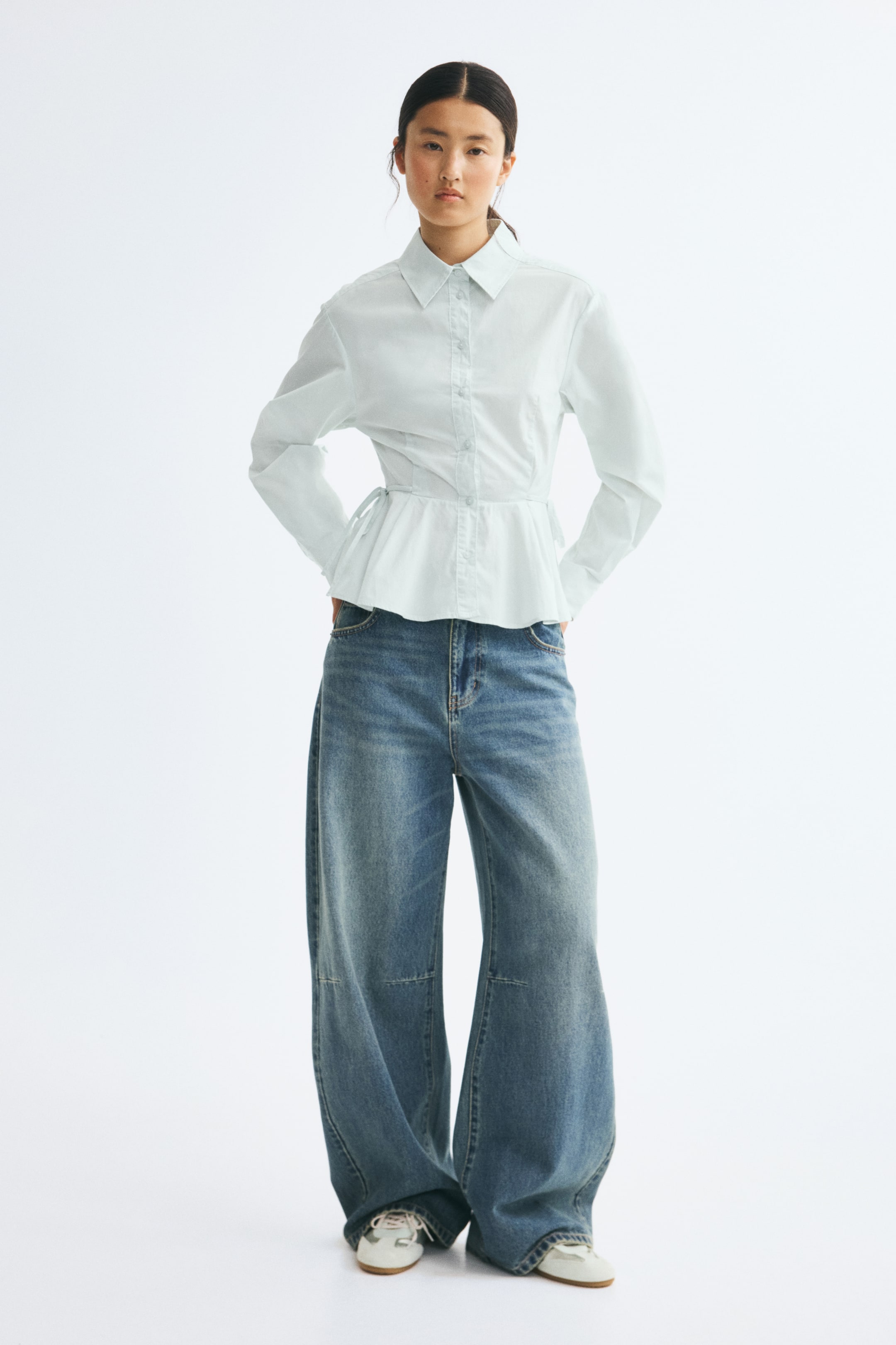 View larger image: A light blue cotton poplin shirt is shown from the front, featuring a pointed collar, a button-down front, and long balloon sleeves with wide cuffs. The shirt is cinched at the waist with narrow side ties and flares into a pleated peplum hem, styled over wide-leg blue jeans.