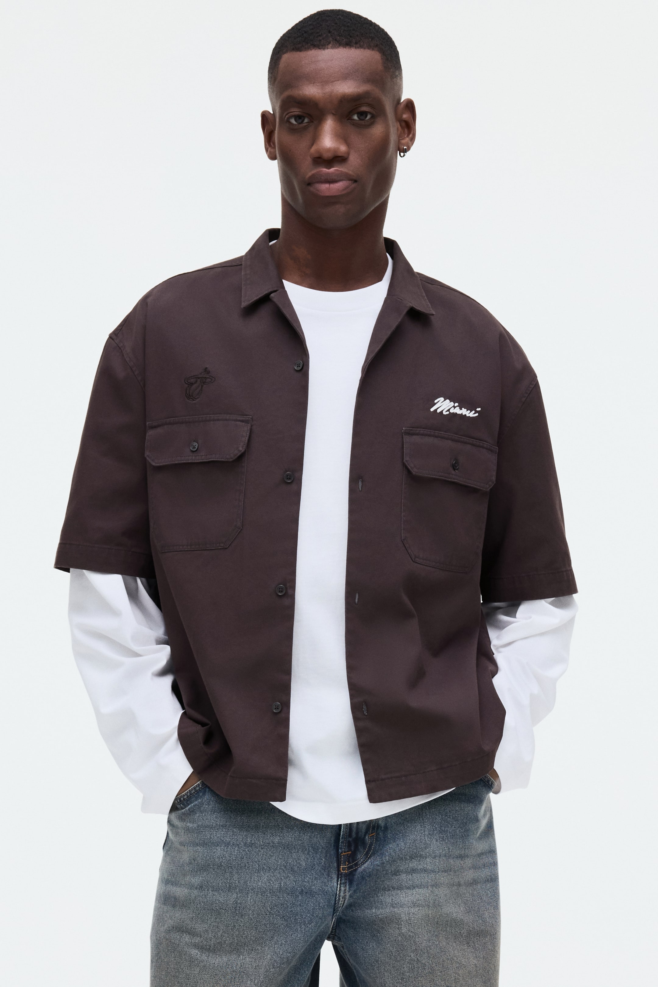 View larger image: A dark brown short-sleeved shirt is showcased from a frontal view, featuring a spread collar and a button-up front closure. The shirt is designed with two buttoned flap pockets on the chest, an embroidered stylized flame on the left, and "Miami" in white script on the right, and is worn open over a white long-sleeved t-shirt.