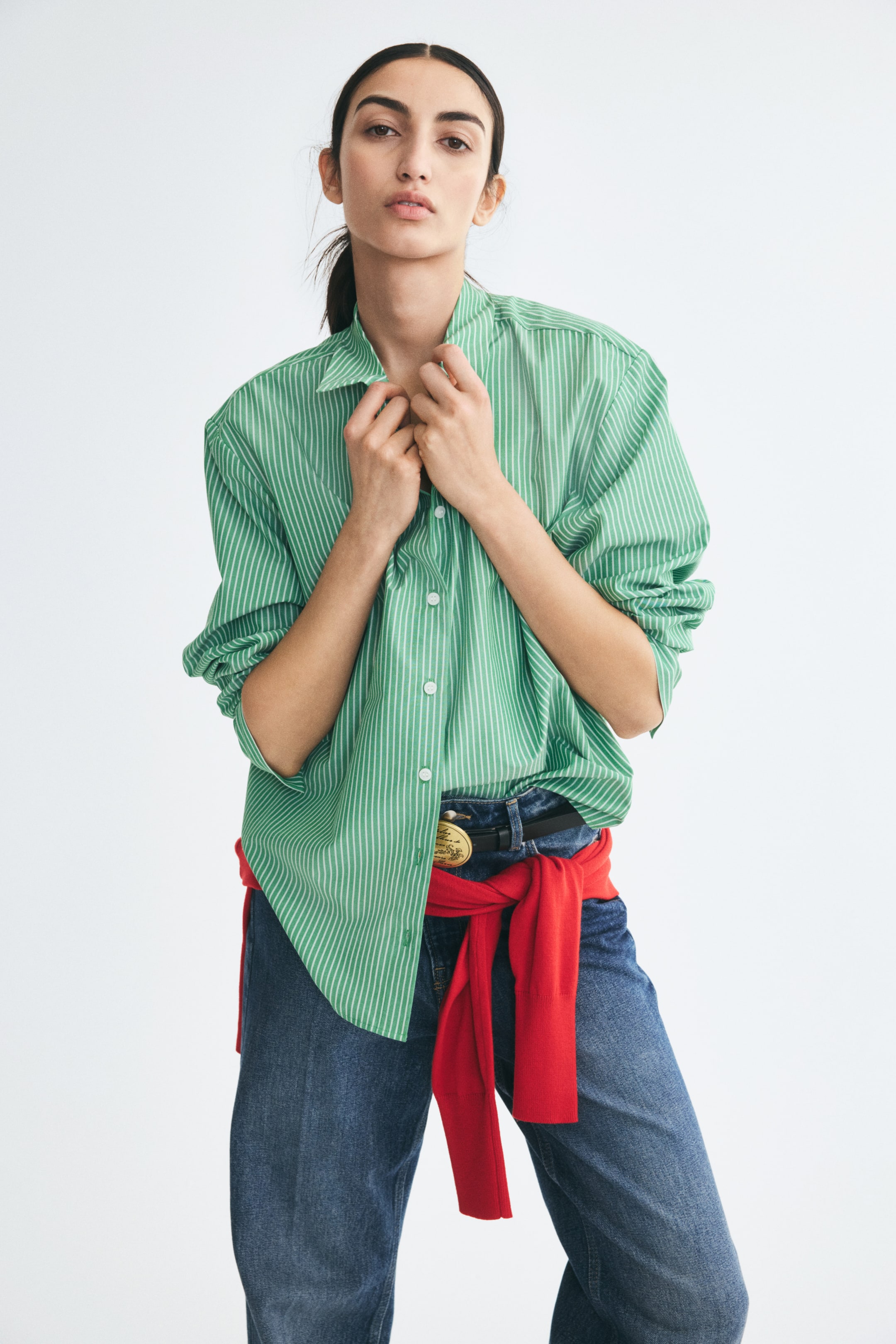 View larger image: A green and white vertically striped woven shirt with a collar and front buttons is worn by the model, who is holding the collar with both hands. The long-sleeved shirt is styled with blue jeans and a red sweater tied around the waist.