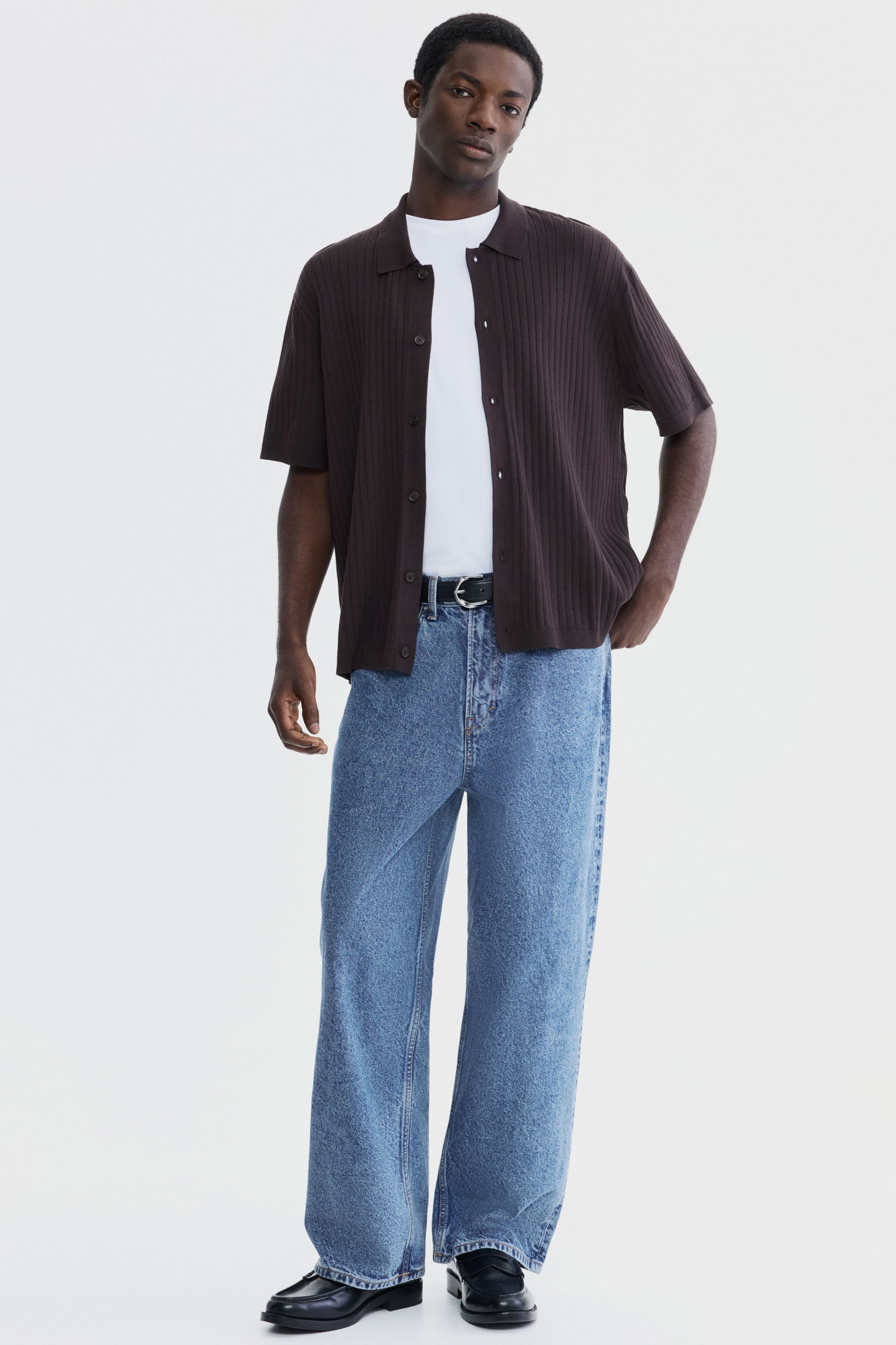 Größeres Bild anzeigen: Die blauen Jeans zeichnen sich durch eine helle Waschung und einen lockeren, weiten Beinabschluss aus, der ihre volle Länge von vorne zeigt. Sie sind hoch tailliert und werden mit einem schwarzen Gürtel gesichert, wobei der Stoff leicht über den Schuhen fällt.