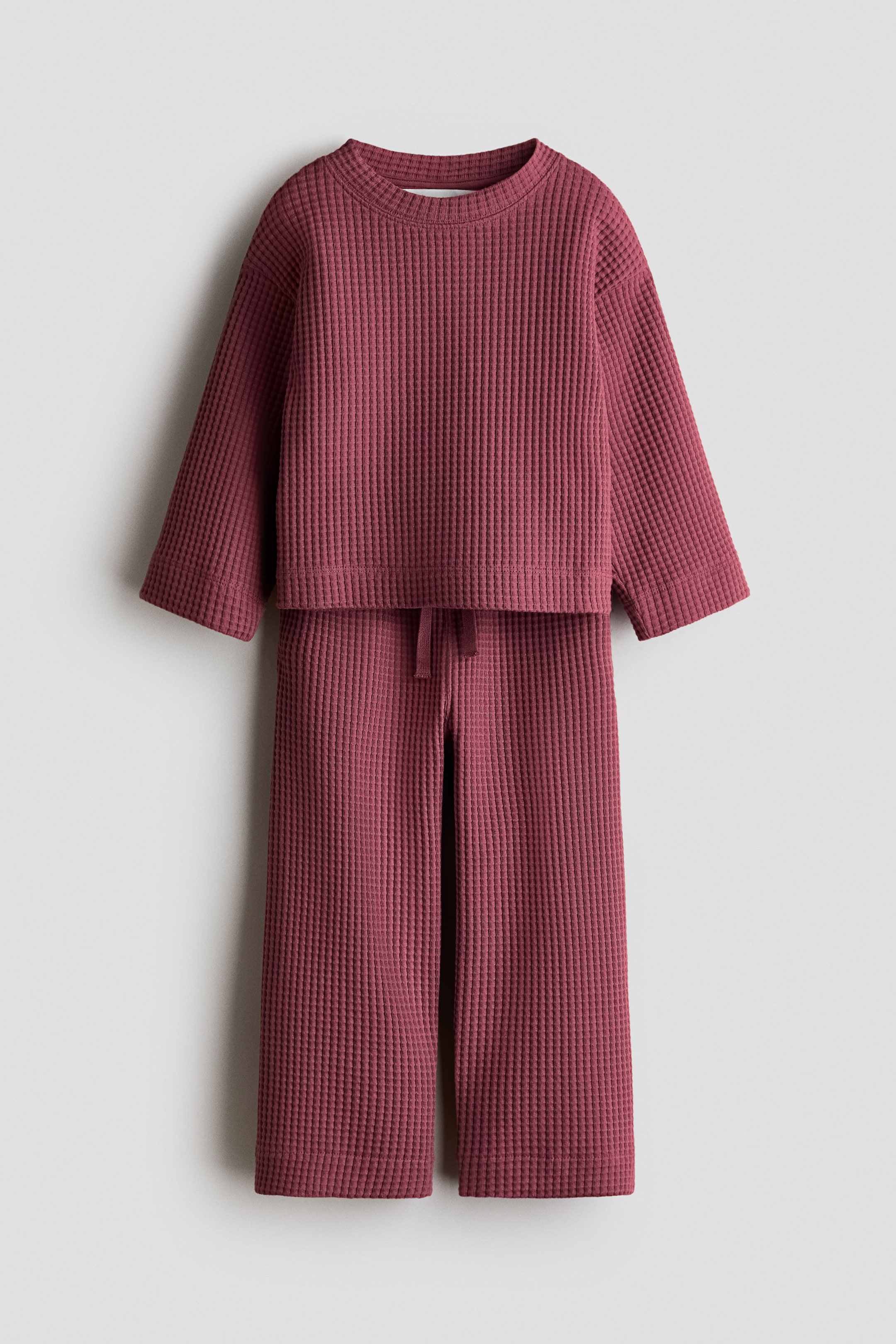 Größeres Bild anzeigen: Das H&M Adorables Set zeigt eine Vorderansicht eines dunkelroten Waffel-Jersey-Oberteils und der dazu passenden Hose. Das Oberteil im Boxy-Stil verfügt über einen runden Halsausschnitt, überschnittene Schultern und lange Ärmel, während die weit geschnittene Hose einen elastischen Bund mit Kordelzug aufweist.