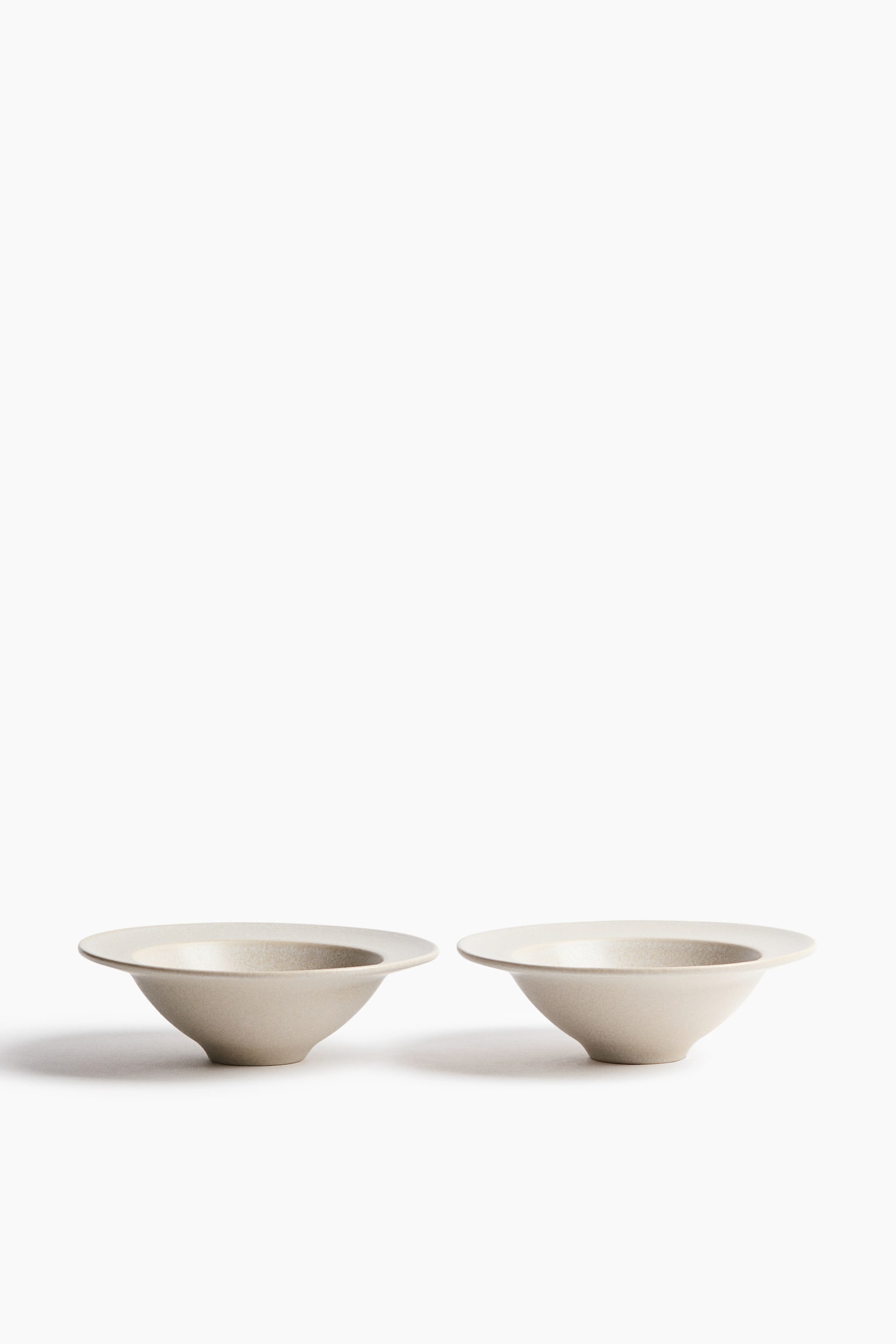 Deux bols en cÃ©ramique blanc cassÃ© sont posÃ©s cÃ´te Ã  cÃ´te, mettant en valeur une forme conique effilÃ©e qui sâÃ©vasent vers un large bord plat. Les bols prÃ©sentent un Ã©maillage mat Ã  la texture fine et sont montrÃ©s de face.