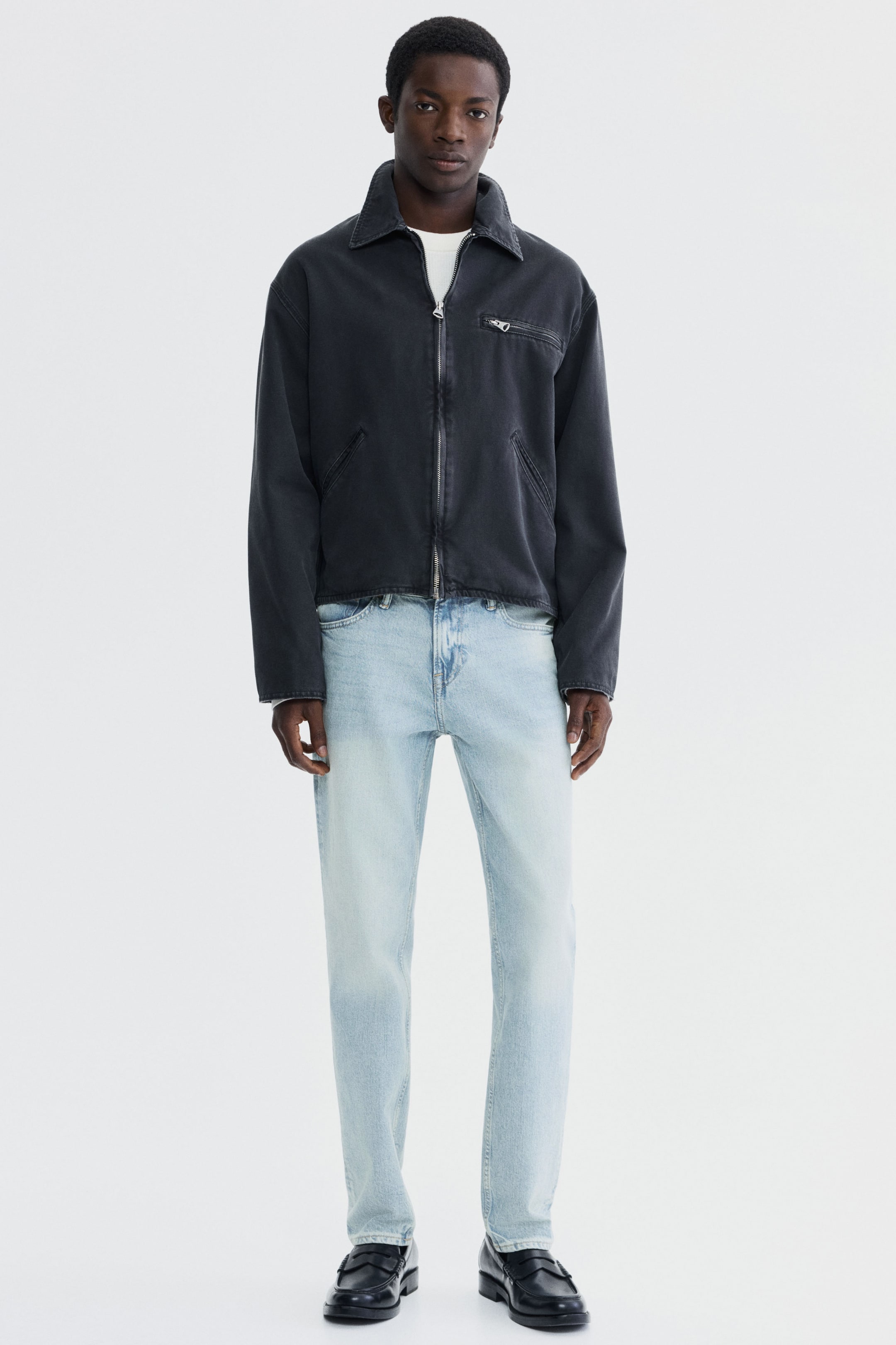 Agrandir l'image: Une veste gris foncé délavée est présentée de face, avec un col pointu, une fermeture éclair argentée sur toute la longueur, deux poches passepoilées inclinées et une poche poitrine zippée, portée sur un t-shirt blanc. Un jean bleu délavé clair avec une coupe droite est porté avec des mocassins noirs.