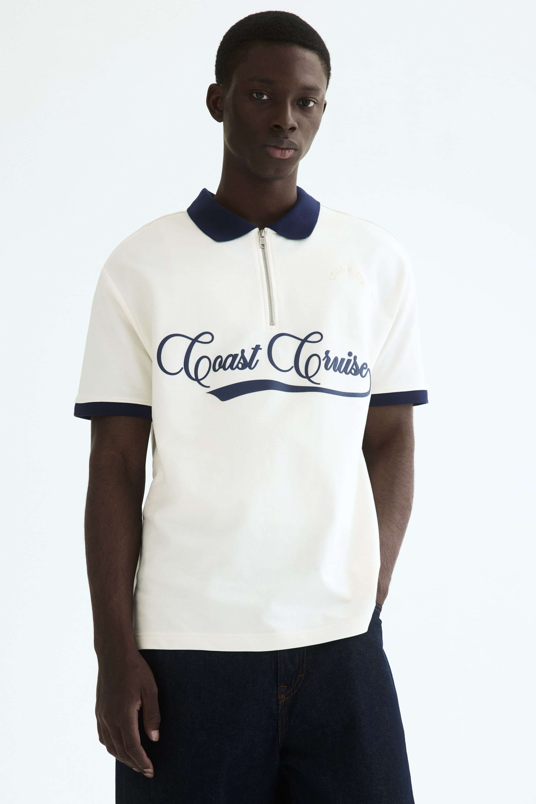 View larger image: A cream short-sleeved polo shirt featuring a navy blue collar and a quarter-zip placket. Navy blue trim borders the sleeve cuffs, and "Coast Cruise" in navy blue script lettering is prominently displayed across the front.