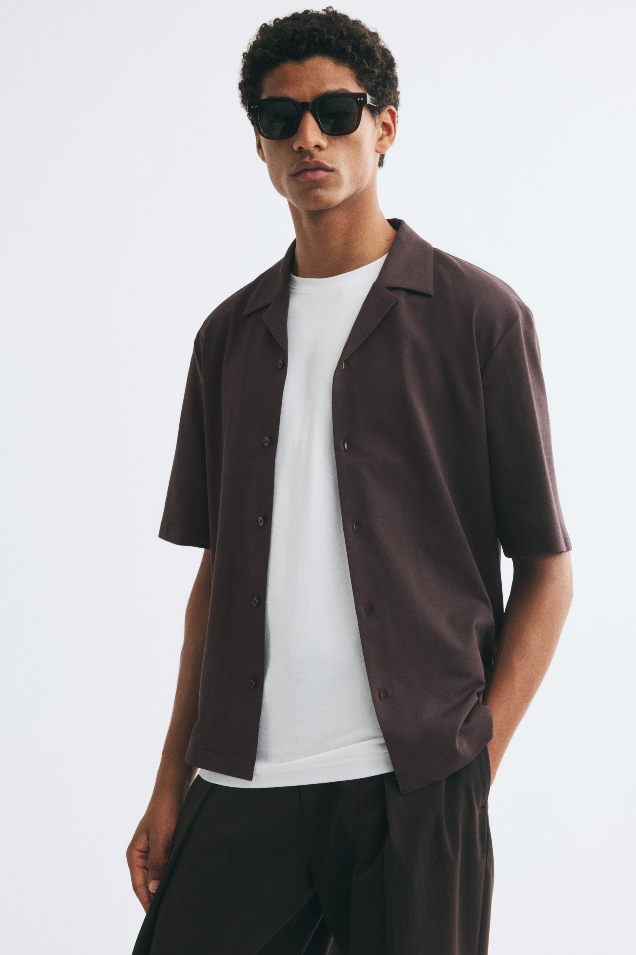 View larger image: A dark brown, short-sleeve jersey shirt with a resort collar is worn open by the model. It is layered over a white crew-neck t-shirt and paired with dark trousers.