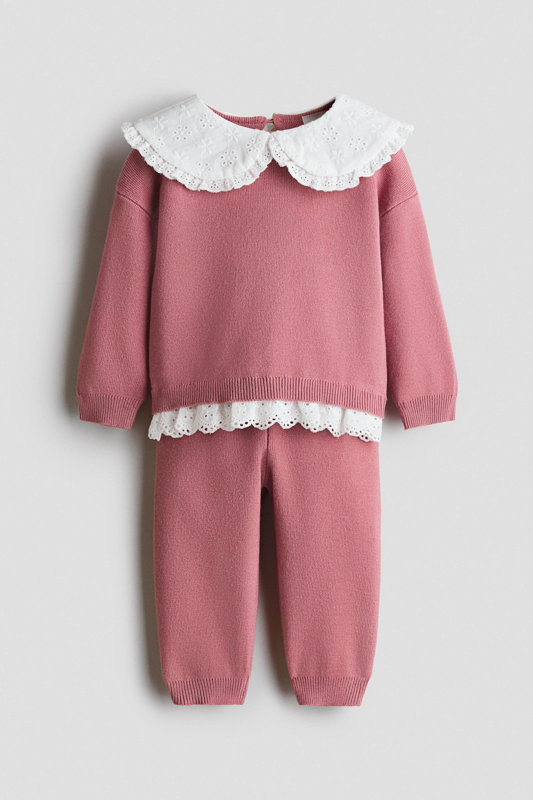 View larger image: A pink knitted two-piece outfit is shown from the front, featuring a sweater with a white eyelet-embroidered Peter Pan collar, ribbed cuffs, and a ribbed hem, with a white eyelet lace trim peeking from underneath. Matching knitted pants complete the set, also featuring ribbed ankle cuffs.