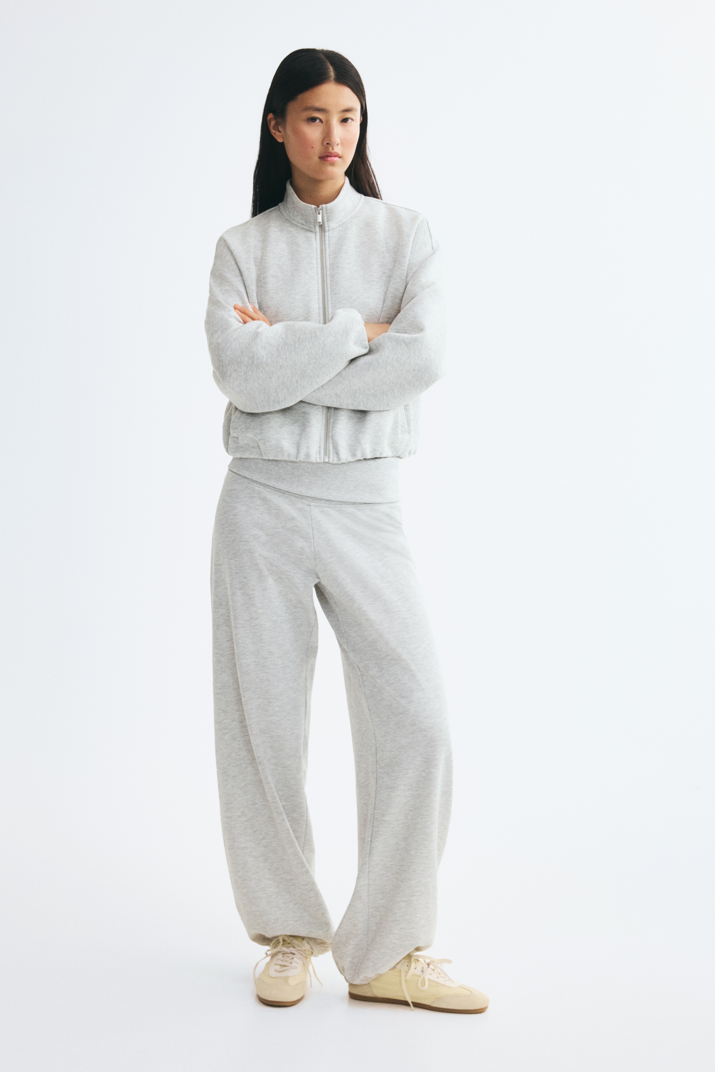 A pair of light grey marl sweatpants is shown from the front, featuring a wide, flat waistband and a relaxed, loose-fitting leg design. The trousers taper down to gathered, elasticated cuffs at the ankles.