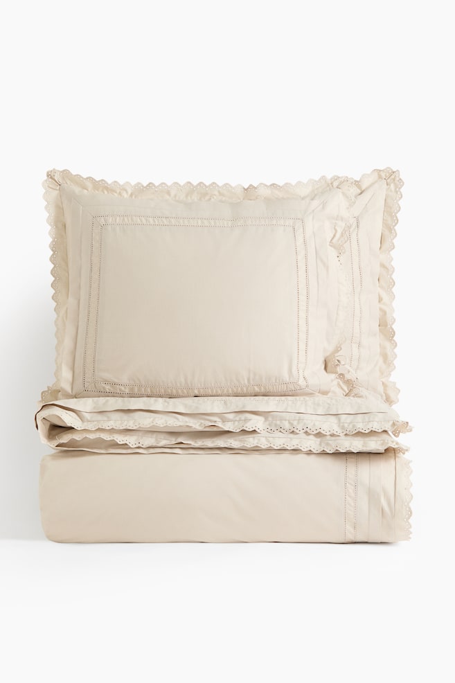 Um conjunto de roupa de cama em tom creme apresenta uma fronha decorativa colocada na vertical sobre uma capa de edredão dobrada, vista de frente. A fronha decorativa destaca-se por uma borda retangular com ponto aberto e um delicado acabamento em renda ajurada com rebordo ondulado nas extremidades, em harmonia com o acabamento e os detalhes de pregas finas na roupa de cama dobrada por baixo. - 1