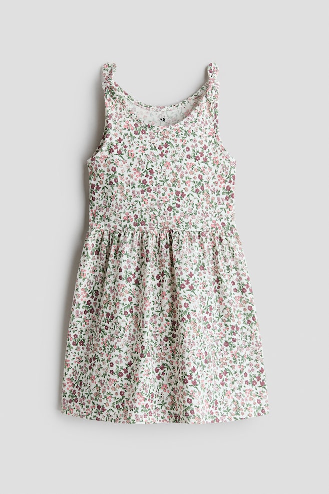 El vestido sin mangas se presenta de frente, confeccionado en un suave jersey de algodón y con un estampado floral completo de pequeñas flores en tonos rosa, verde y granate sobre un fondo blanco. Está diseñado con un escote redondo, detalles de nudos decorativos en los hombros, una costura fruncida en la cintura que añade volumen y una falda ligeramente acampanada. - 1