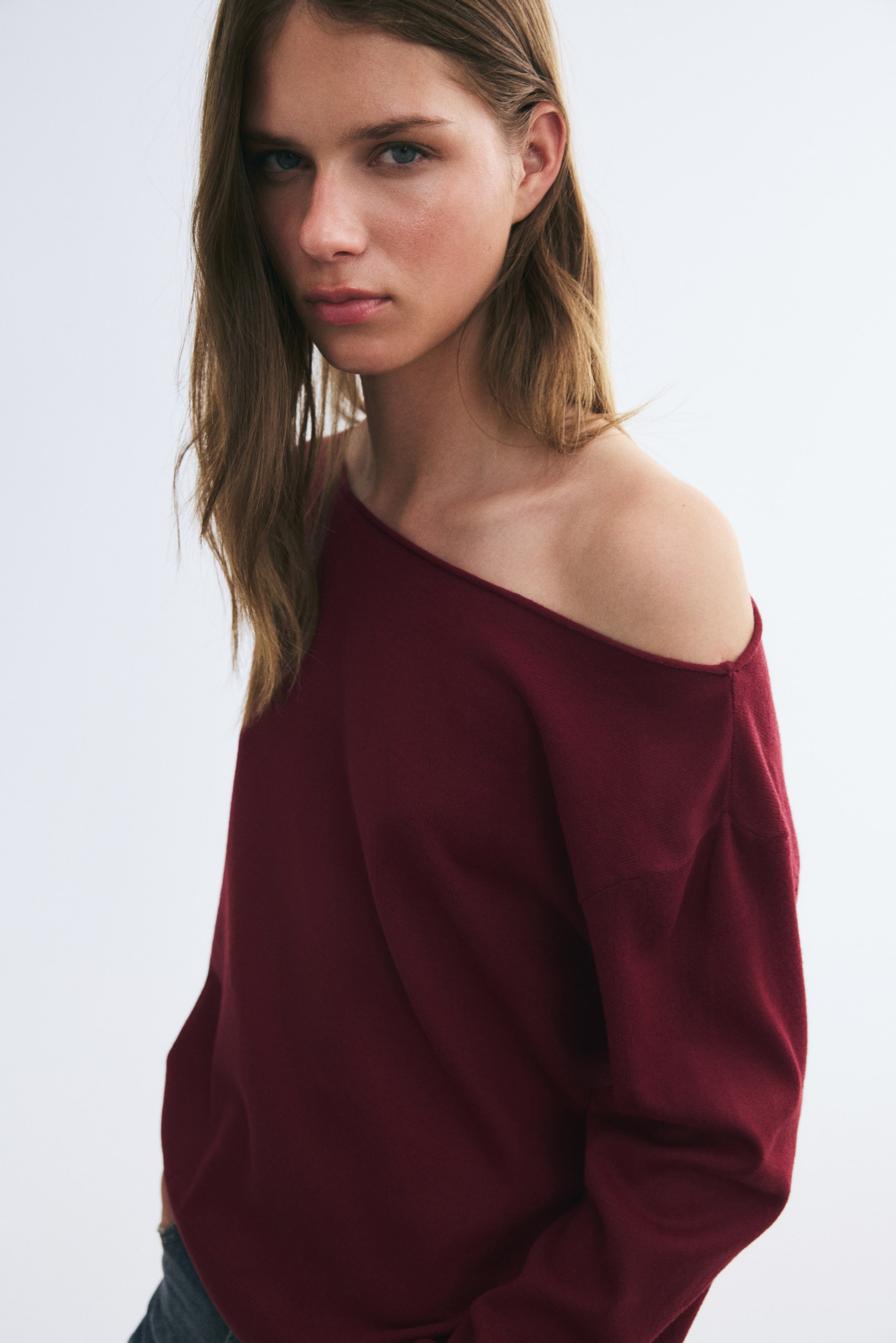 View larger image: A burgundy off-the-shoulder sweater in a soft knit is being worn by the model, featuring a roll-edge neckline and dropped shoulders. The sweater is draping off one shoulder, showing its relaxed fit.