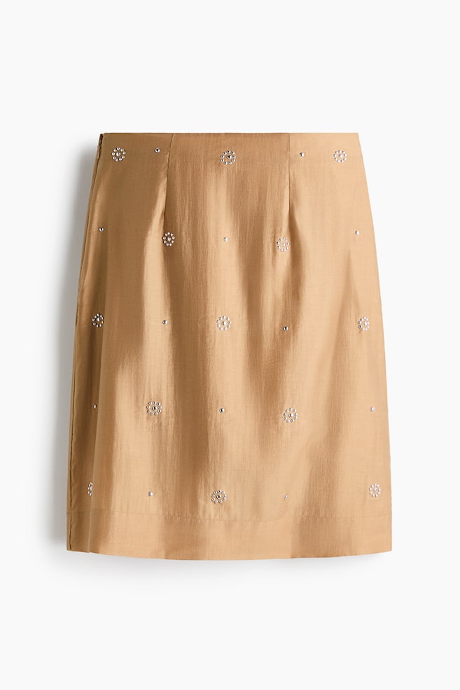 A knee-length skirt in a tan lyocell-blend weave is shown from the front, featuring a subtle sheen and a structured waist with darts. The skirt is adorned with all-over silver-toned stud embellishments arranged in alternating floral patterns and single dots. - 1