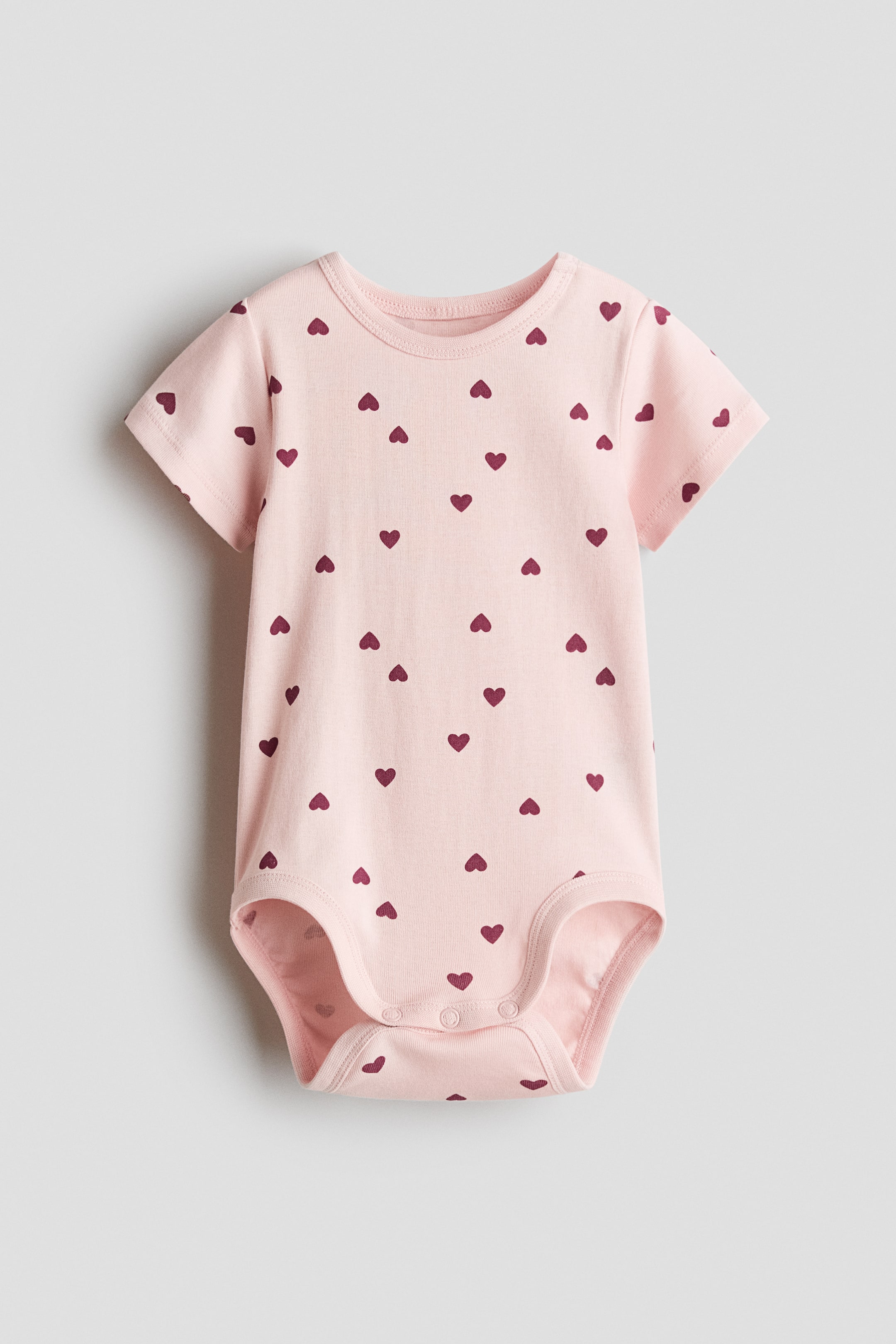 View larger image: A short-sleeved bodysuit in soft light pink cotton jersey showcases an all-over print of small dark red hearts. It features a round, trimmed neckline and press-studs on the left shoulder and at the bottom.