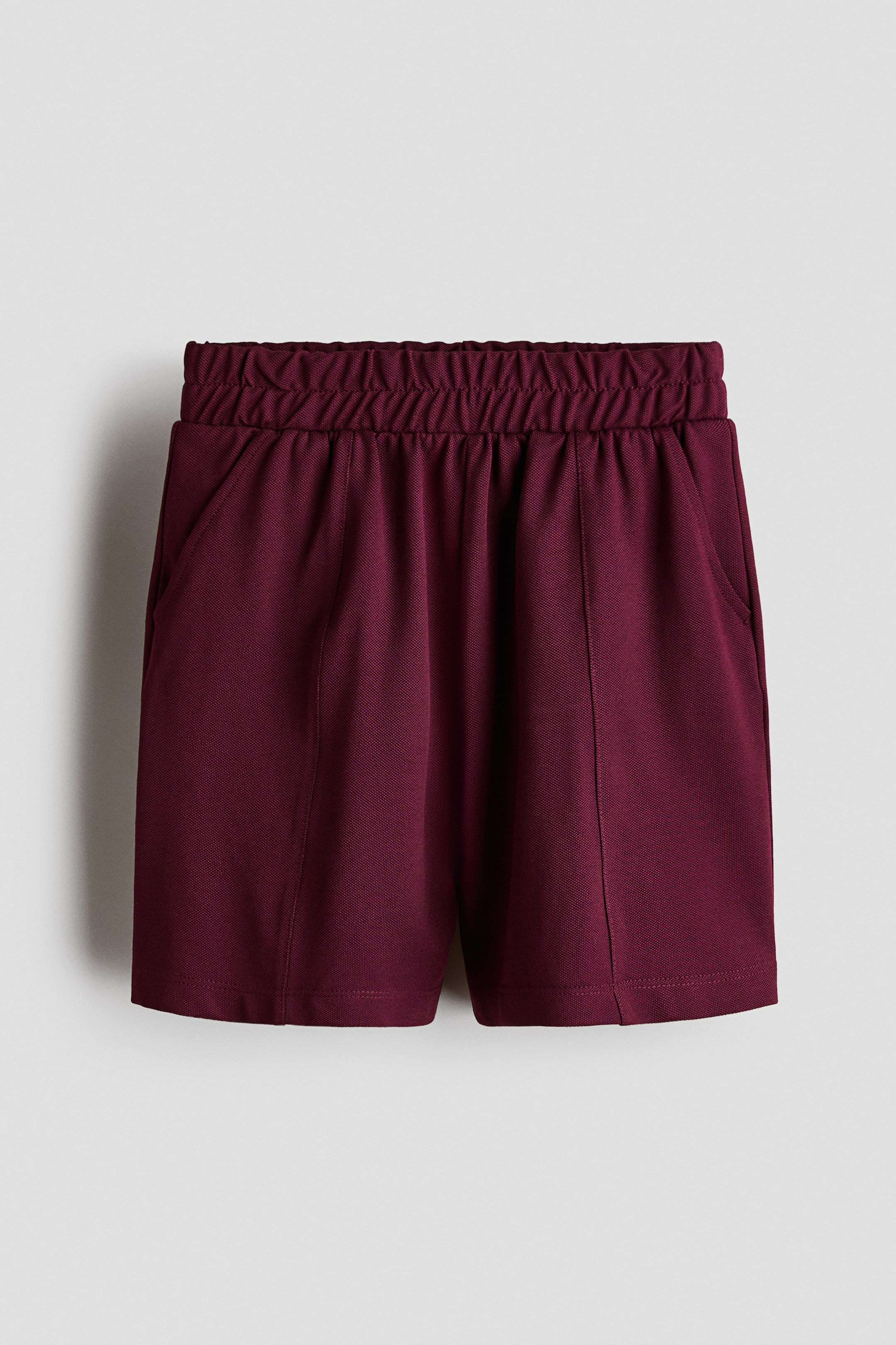 Xem ảnh lớn hơn: Một chiếc quần short màu đỏ burgundy được chụp từ phía trước, nổi bật với cạp chun bản rộng và phom dáng thoải mái. Quần short có các đường may pintuck dọc chạy xuống mặt trước của mỗi ống quần và túi bên xéo.
