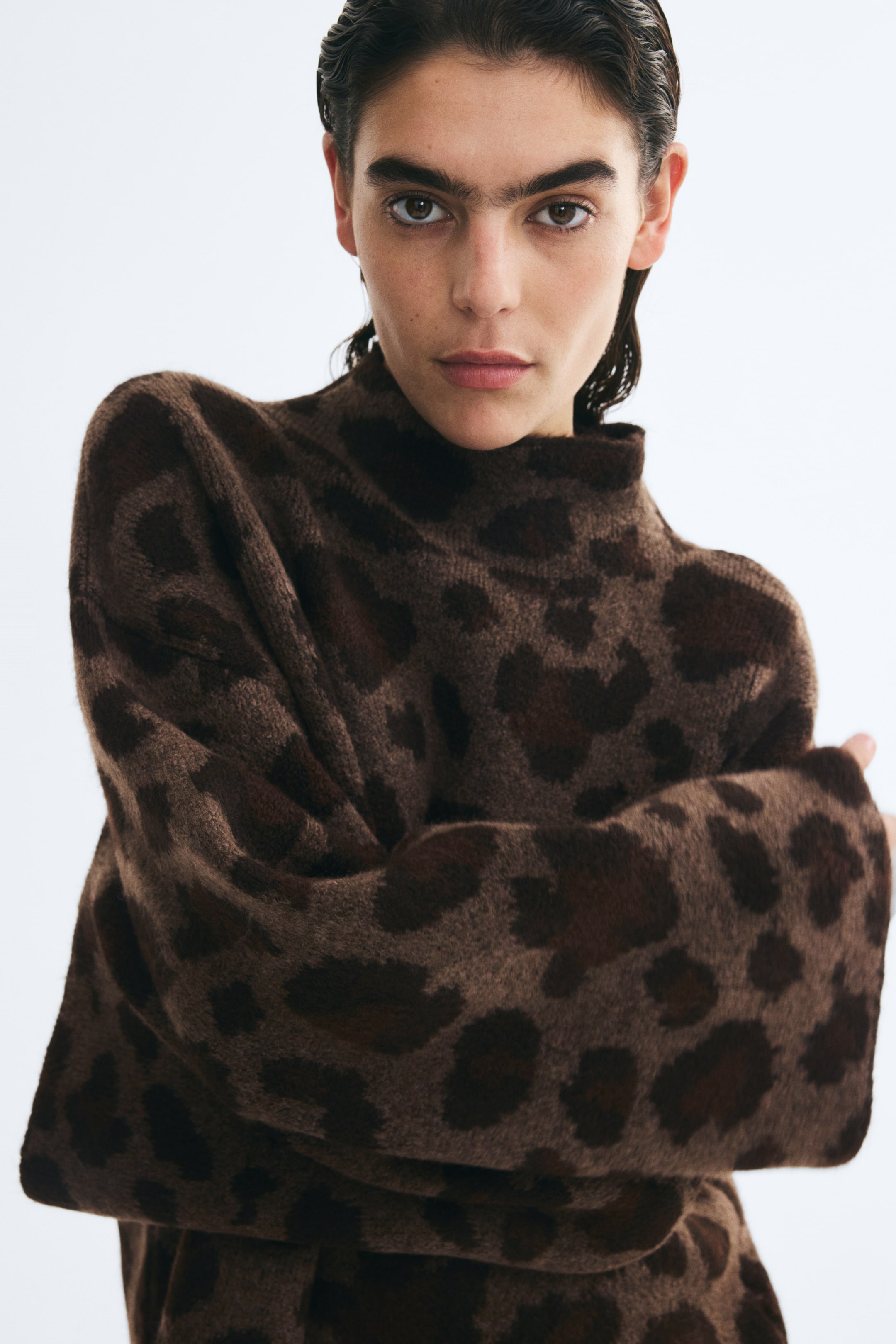 Se større billede: En brun jacquardstrikket sweater med leopardprint, høj hals og sænkede skuldre bæres af modellen. Modellen krydser armene og fremhæver beklædningsgenstandens bløde tekstur samt de lange, brede ærmer.