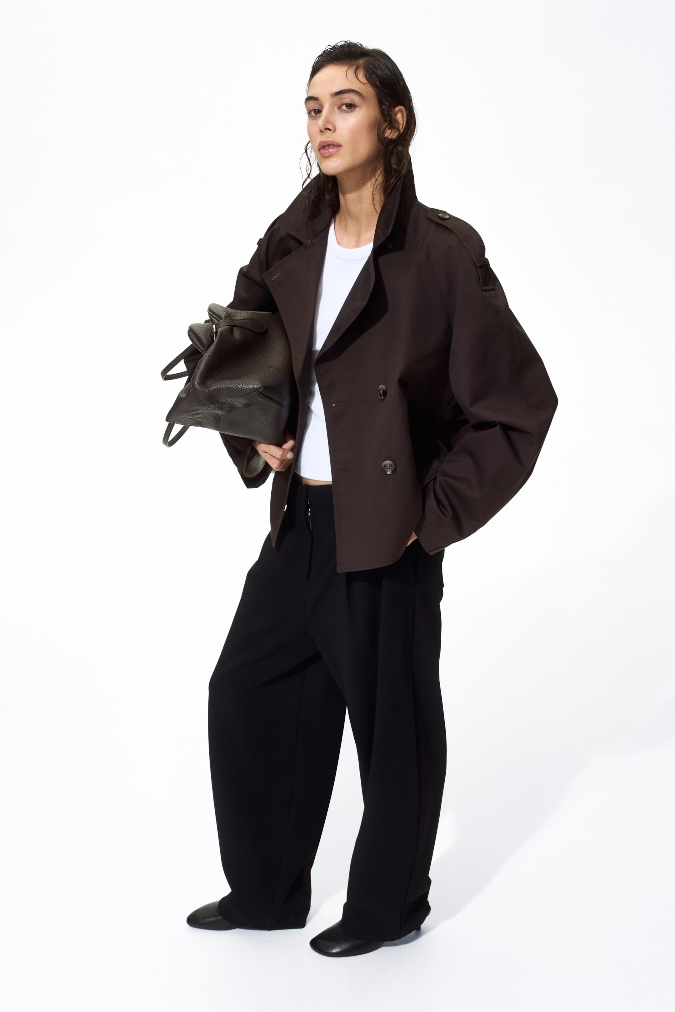 Ingrandisci l'immagine: Una giacca corta marrone scuro è presentata da un angolo leggermente frontale, caratterizzata da un design doppiopetto con due file di bottoni, un ampio colletto, spalline sulle spalle e maniche larghe con una vestibilità rilassata. È stratificata sopra una t-shirt bianca.