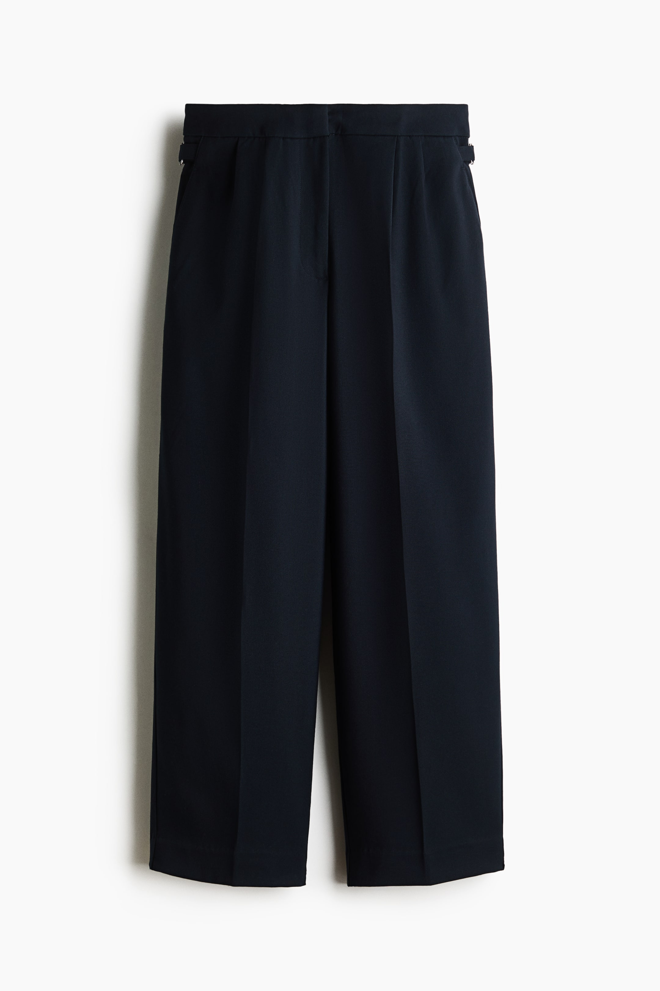 View larger image: A front view of high-waisted, dark navy woven pants featuring straight legs with sharp front creases and pleats below the waistband. The trousers are displaying small adjustable tabs with metal buckles on the sides of the waistband and diagonal side pockets.