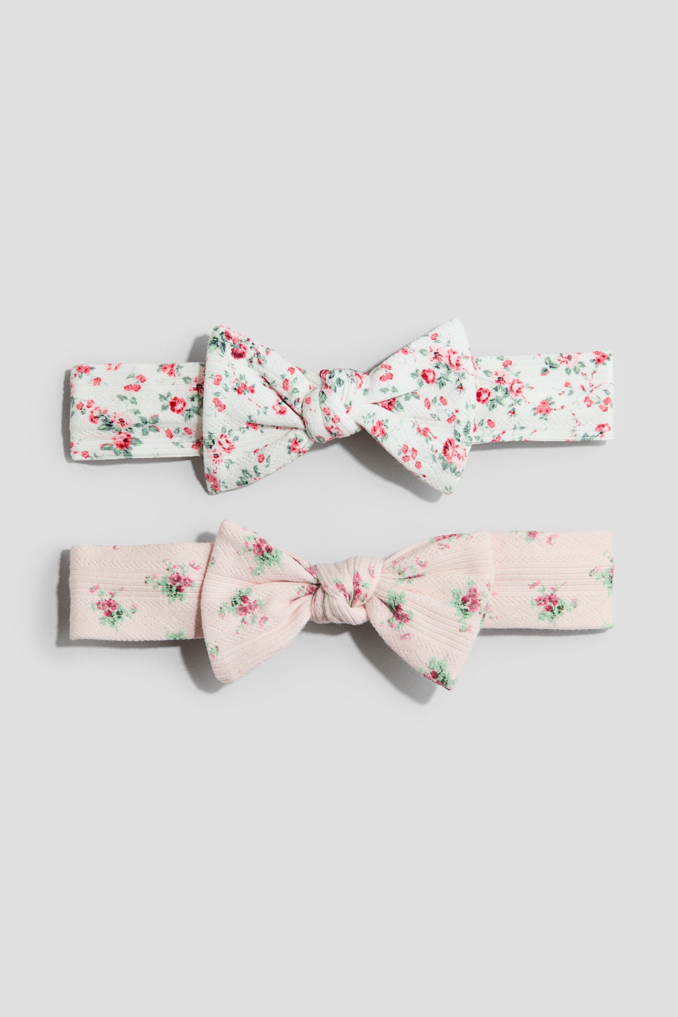 View larger image: Dalawang headbands na may bow detail sa harap, bawat isa ay may banayad na ribbed texture at floral pattern. Ang nasa itaas na headband ay puti na may pulang at berdeng bulaklak, at ang nasa ibabang headband ay mapusyaw na rosas na may rosas at berdeng bulaklak.