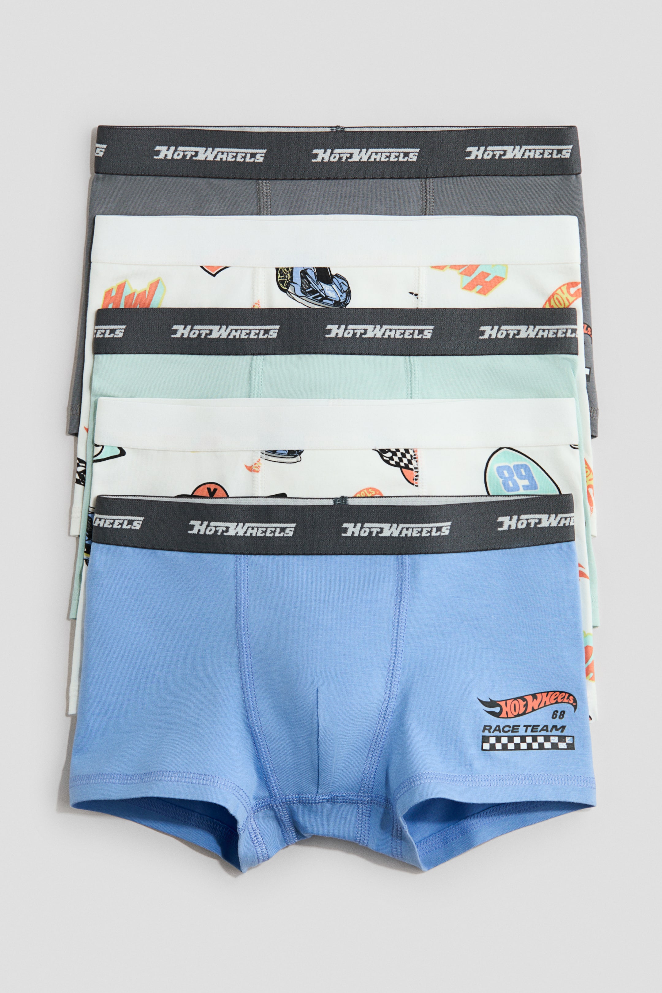 Grösseres Bild ansehen: Fünf Paar Baumwolljersey-Trunks sind gestapelt und zeigen die Vorderseite. Jeder Trunk hat kurze Beine, einen elastischen Bund mit dem "Hot Wheels"-Logo und eine gefütterte Vorderseite. Der Stapel enthält Paare in Dunkelgrau, Hellgrün, Hellblau mit einem "Hot Wheels Race Team"-Logo und zwei Paare mit bunten Auto- und Rennflaggen-Prints.