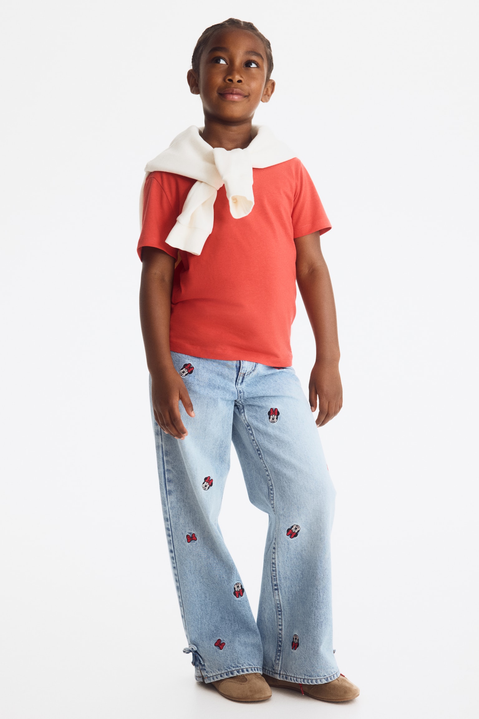 Light blue wide-leg denim jeans are laid flat, showcasing a front view, adorned with embroidered Minnie Mouse heads and red bows scattered across both legs. Small denim bows fasten side slits at the leg openings. - 1