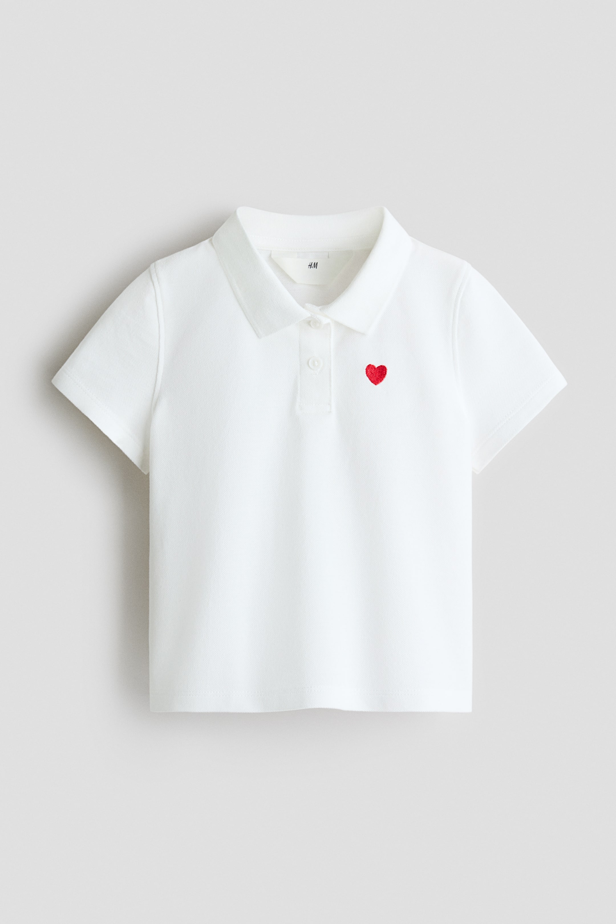 View larger image: A white polo shirt is presented FRONT, featuring a polo collar, short sleeves, and a two-button placket with white buttons. A small red embroidered heart is visible on the left chest.