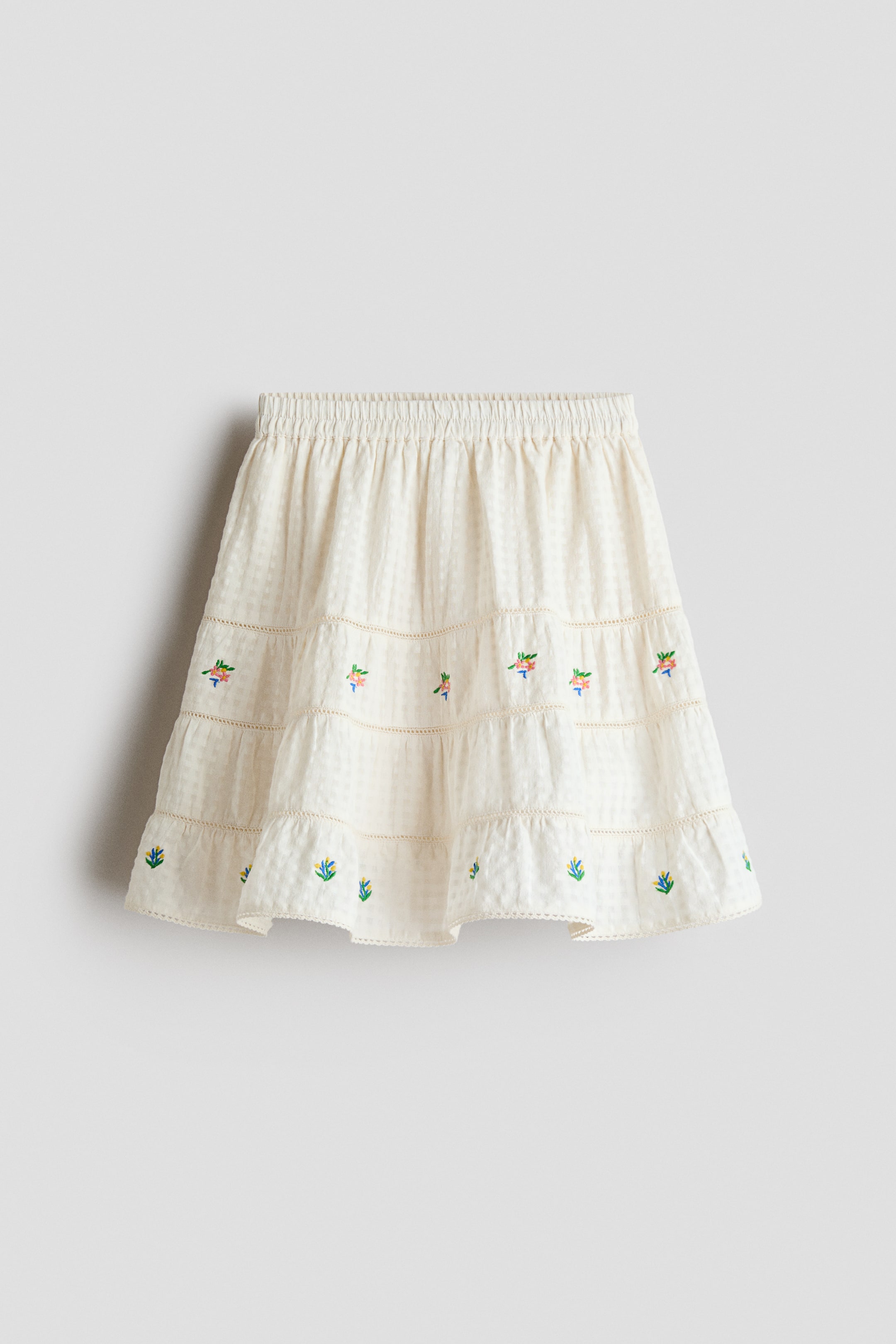 View larger image: An off-white tiered mini-skirt featuring an elastic waistband and a subtle checkered fabric weave. Each of its three gathered tiers showcases small colorful floral embroidery, and the hemline finishes with a delicate scalloped edge.