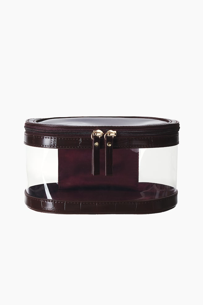 Une trousse de maquillage ovale est prÃ©sentÃ©e de face, avec un corps en plastique transparent et une finition faÃ§on crocodile marron foncÃ© sur le haut et le bas. La trousse est dotÃ©e dâune fermeture zippÃ©e sur tout le pourtour du couvercle, rehaussÃ©e de deux fermetures Ã©clair dorÃ©es assorties Ã  des tirettes marron foncÃ©. - 1