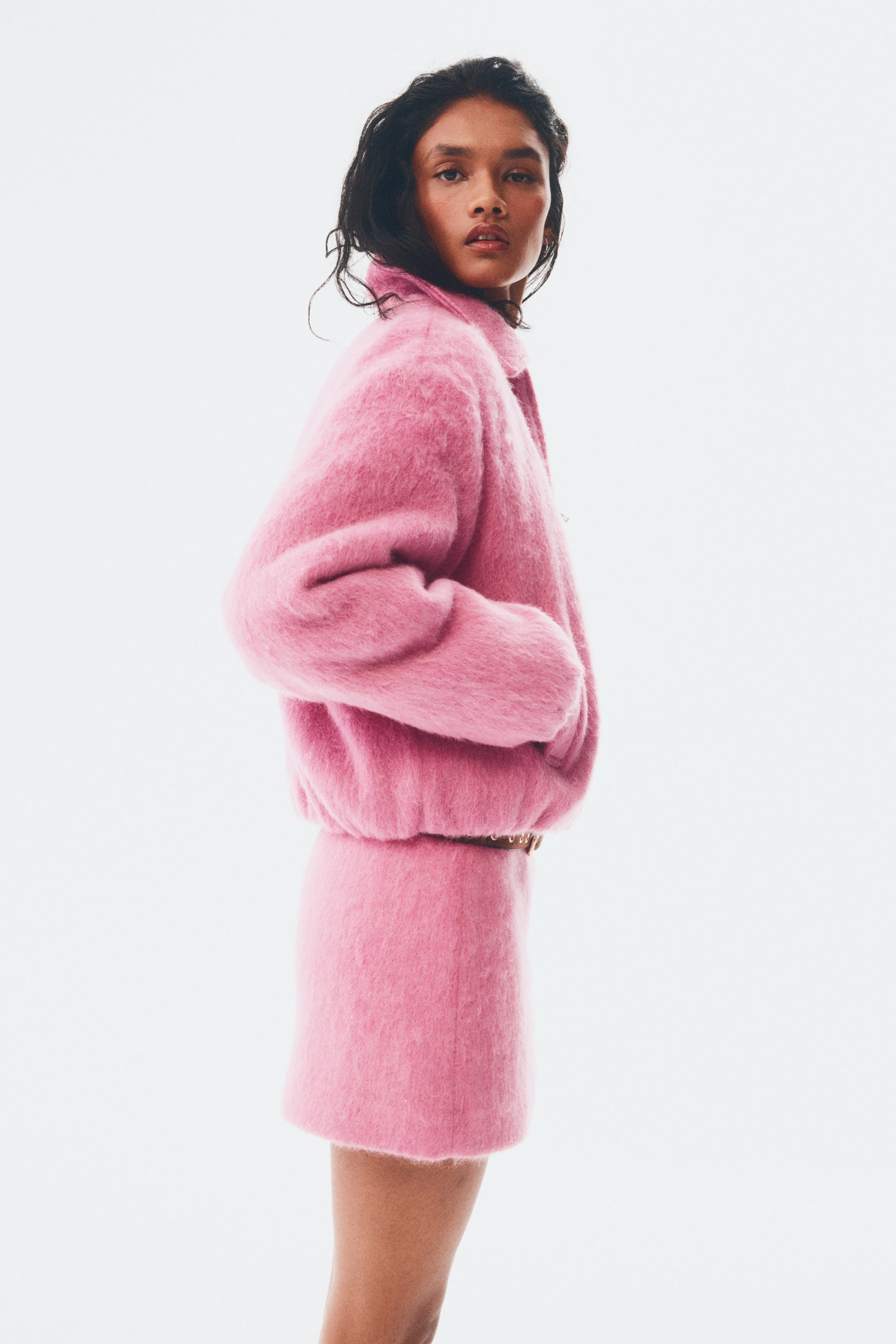View larger image: A pink fuzzy jacket and matching straight-cut mini skirt ensemble is being presented, facing left at a slight angle. The jacket is designed with a pointed collar, full long sleeves, and an elasticated hem that is creating a blouson silhouette, with a thin brown belt visible between the jacket and skirt.