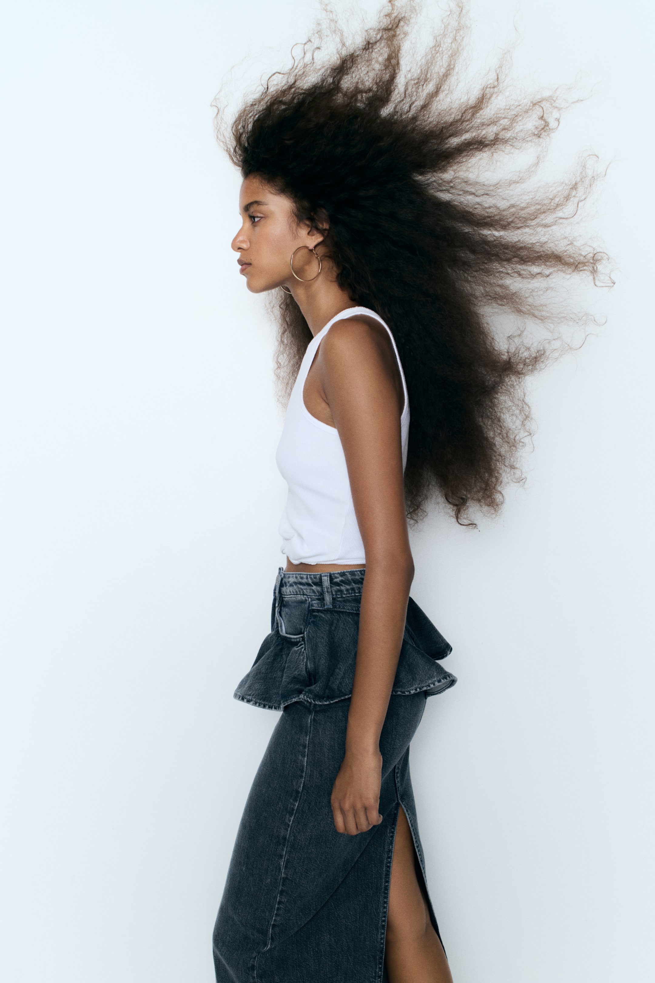 View larger image: A long, fitted denim skirt in a dark grey wash, featuring a wide flounce at the waist and a high back slit, is worn by the model. The skirt is paired with a white sleeveless top.