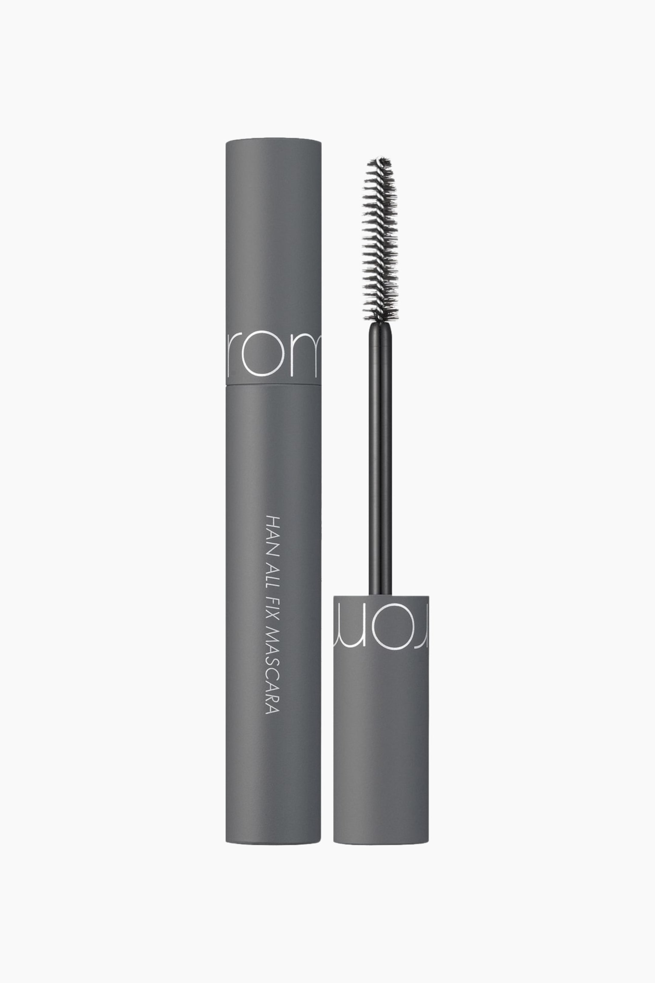 Vis større bilde: En mørk grå mascara-tube står loddrett, med «rom&nd» synlig horisontalt nær toppen og «HAN ALL FIX MASCARA» synlig vertikalt langs siden. Korken, med den svarte mascara-applikatoren festet, er tatt av og plassert til høyre, med børsten rettet oppover og bustkosten godt synlig.