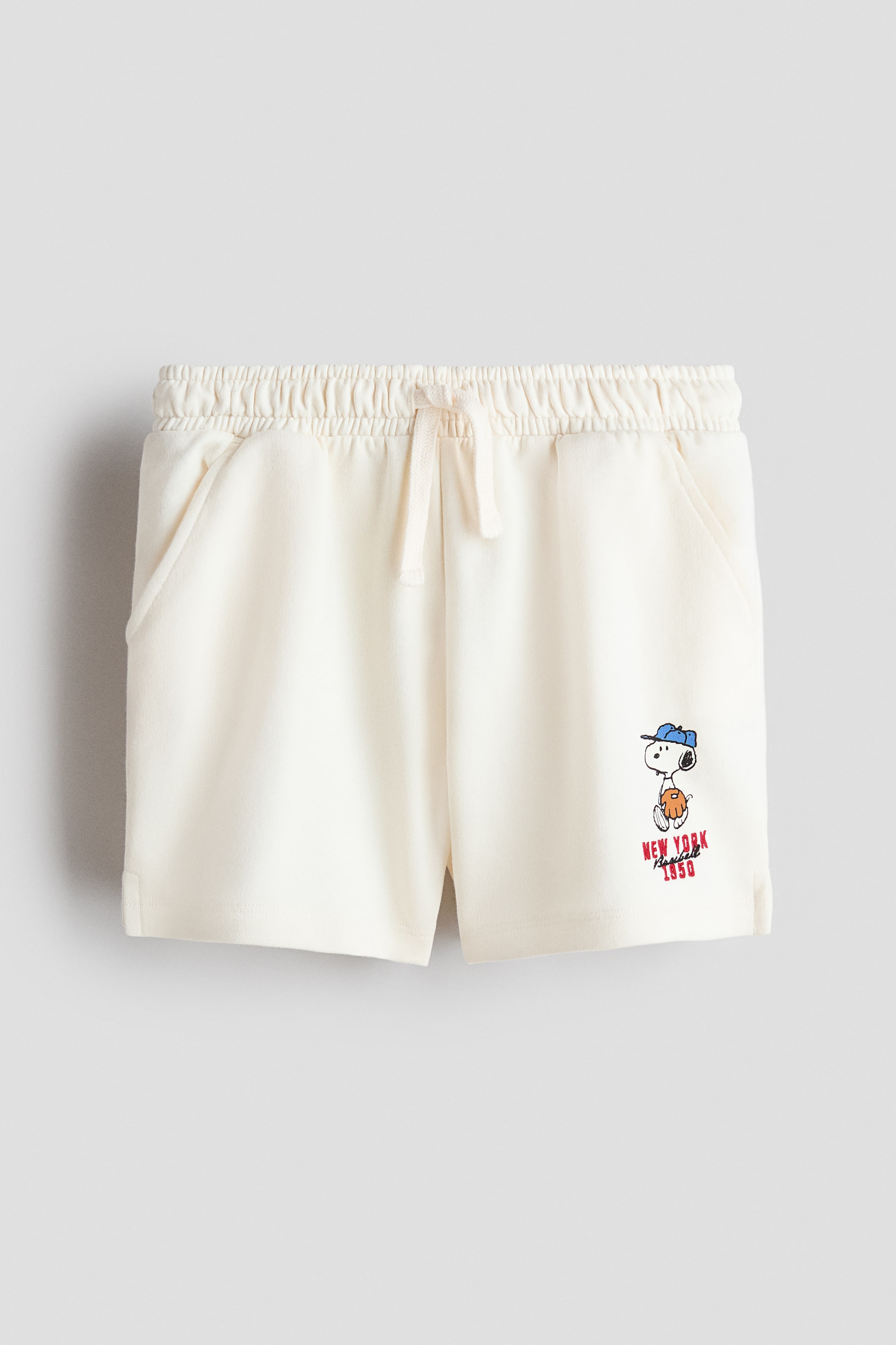 Agrandir l'image: Shorts écru présentés de face, dotés d’une taille élastiquée avec un cordon de serrage crème et de deux poches latérales obliques. Un motif de Snoopy portant une casquette de baseball bleue et un gant apparaît sur la partie basse de la jambe droite, avec, en dessous, un texte rouge indiquant "NEW YORK BASEBALL 1950".