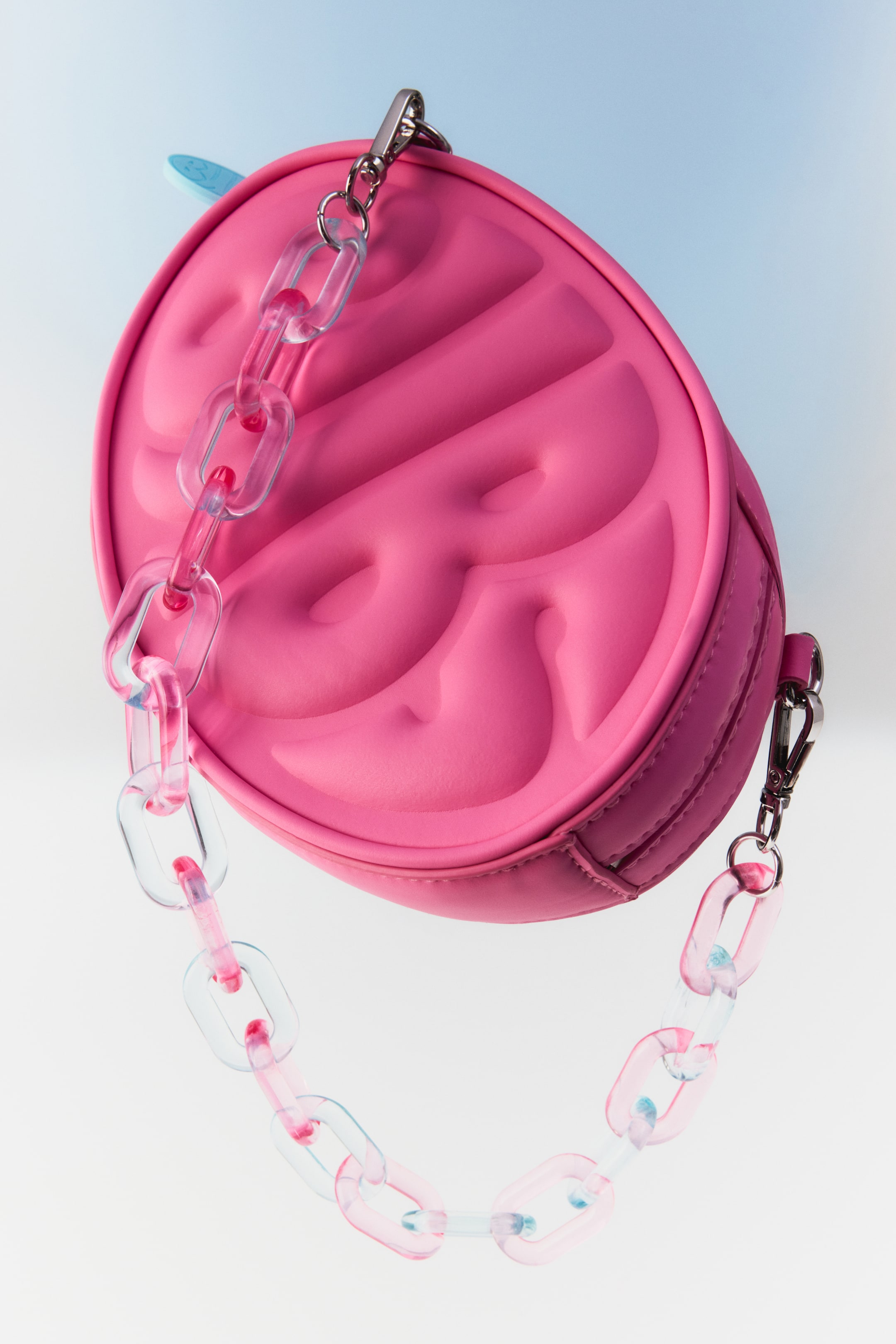 Agrandir l'image: Un sac à main en forme de demi-lune d'un rose éclatant présente son devant, qui est embossé avec des motifs arrondis répétés. Une bandoulière en chaîne épaisse et translucide, avec des maillons alternant entre transparent et teinté de rose, se fixe au sac avec des mousquetons de couleur argentée.