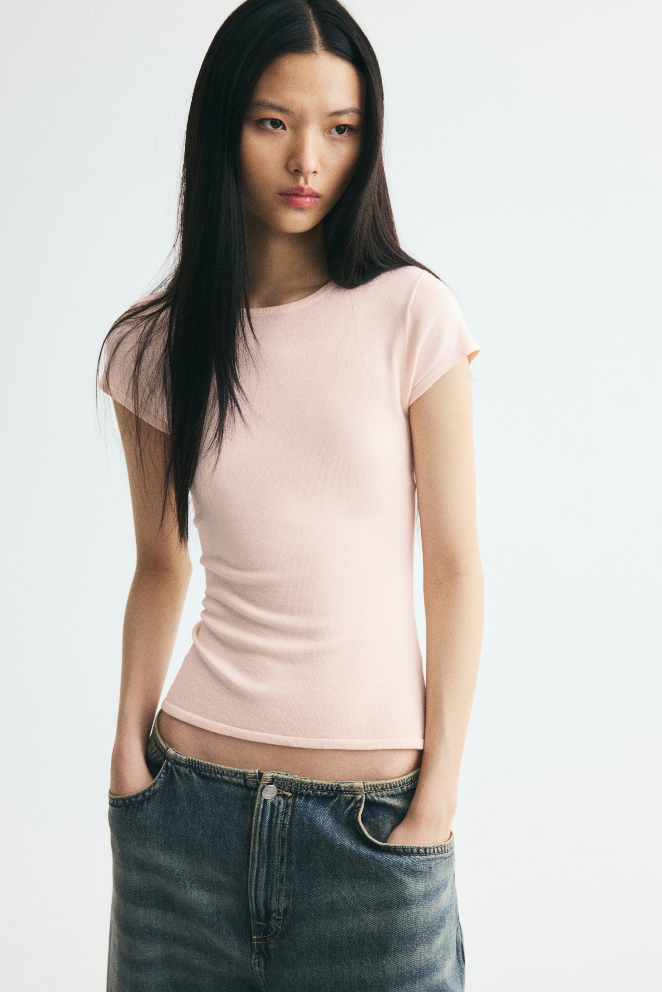 View larger image: The model is wearing a light pink, fitted fine-knit top featuring a round neckline and cap sleeves. The top is styled with low-rise blue jeans, with the model resting her hands in the front pockets.