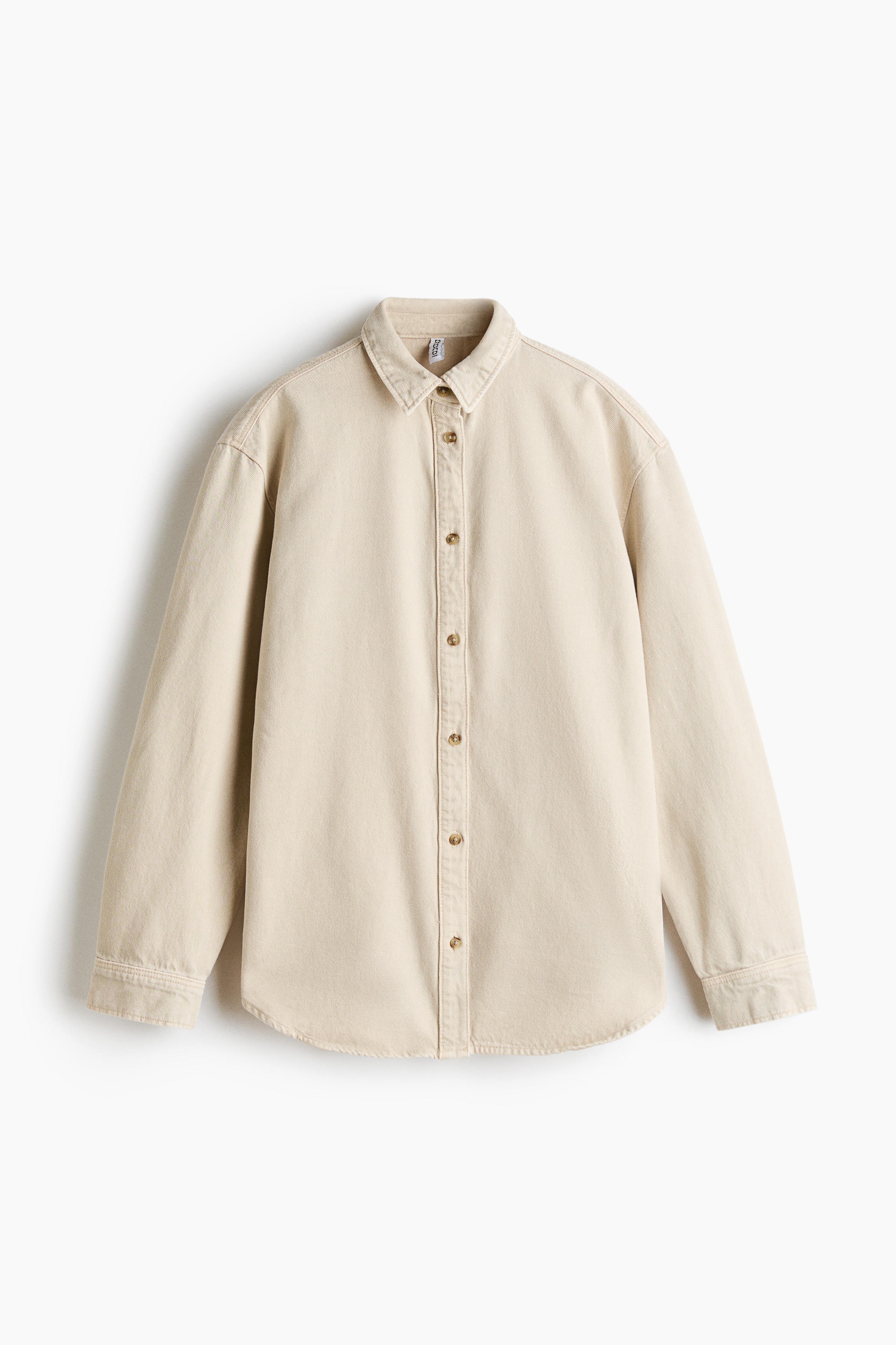 Een oversized lichtbeige denimhemd wordt open gedragen door het model, dat gehurkt op een witte metalen kruk zit. Het hemd heeft een kraag, een sluiting met knopen vooraan, verlaagde schoudernaden en lange mouwen, en is gestyled over een wit T-shirt en een bijpassende lichtgekleurde jeans.