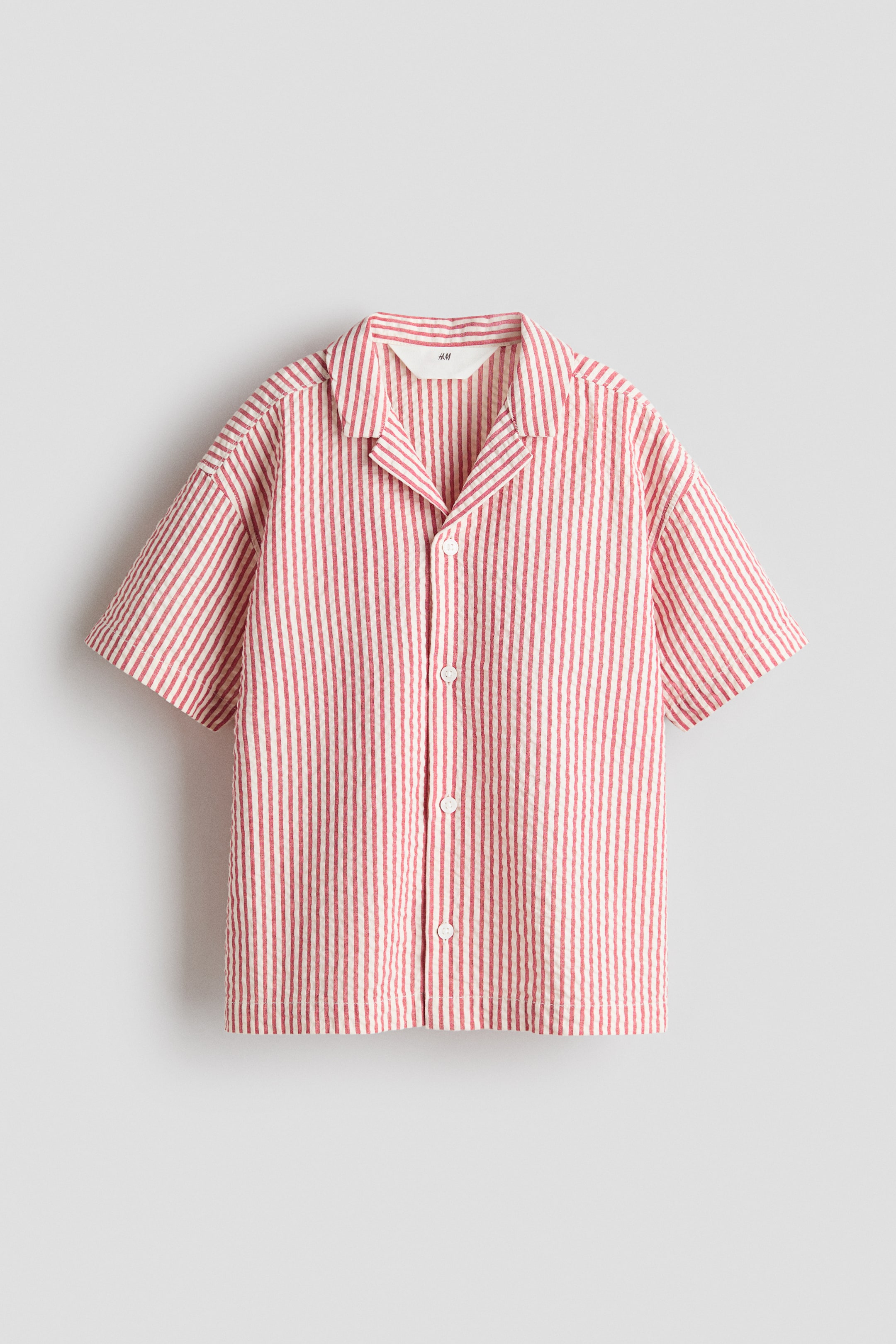 View larger image: A red and white striped seersucker short-sleeved shirt is shown from the front, featuring a camp collar and white buttons down the centre.
