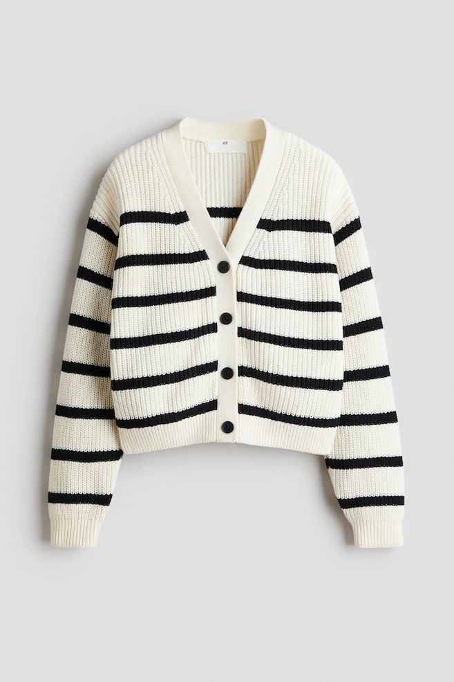 Un cardigan a coste a righe crema e nero è mostrato frontalmente, caratterizzato da uno scollo a V e cinque bottoni neri che corrono lungo il centro. È progettato con spalle cadenti e coste lungo l'abbottonatura, i polsini e l'orlo. - 1