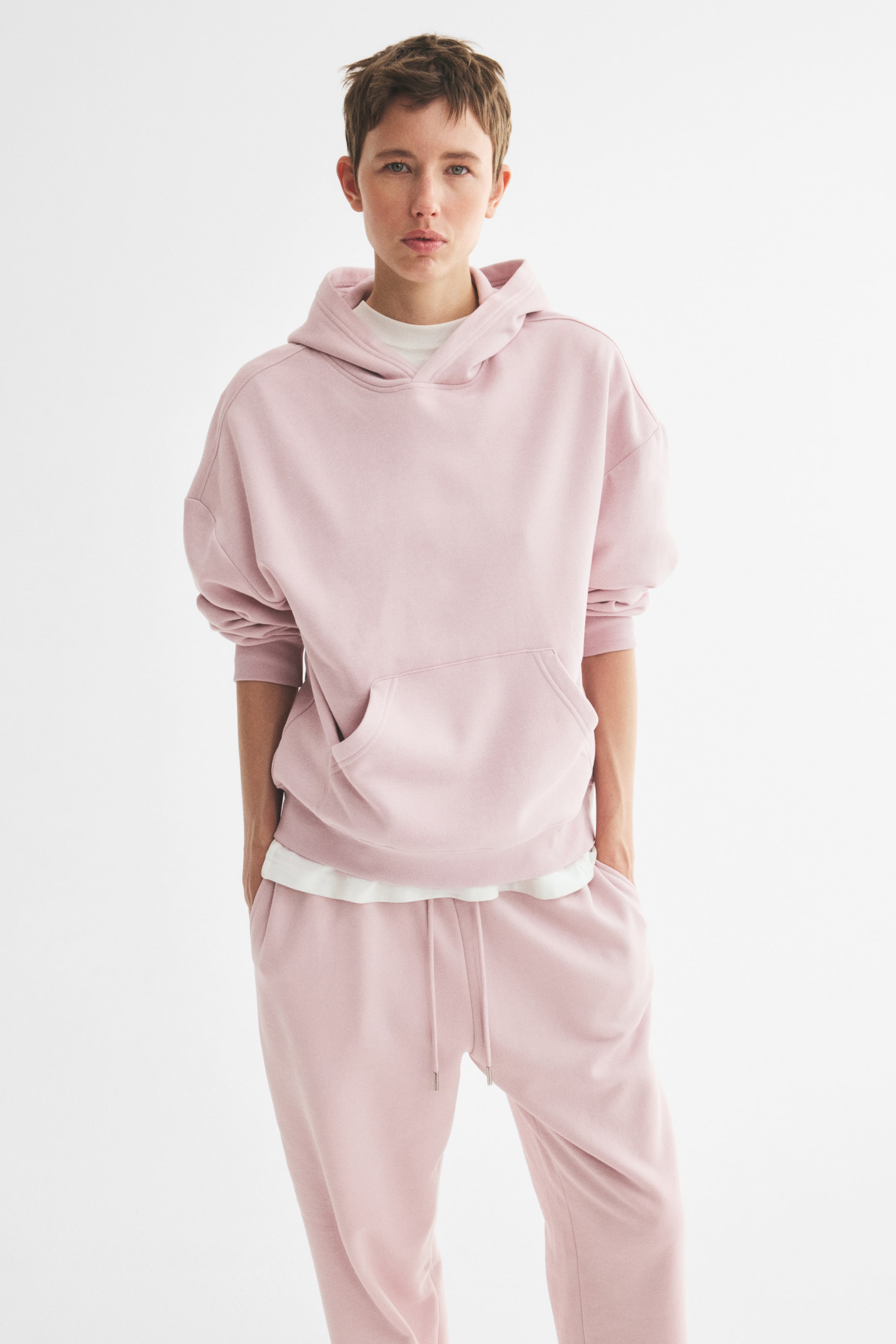 View larger image: A pink hoodie with a double-layered hood, low dropped shoulders, a kangaroo pocket, and ribbing at the cuffs and hem is worn by the model. A white t-shirt is layered underneath, and the model places their hands into the kangaroo pocket.