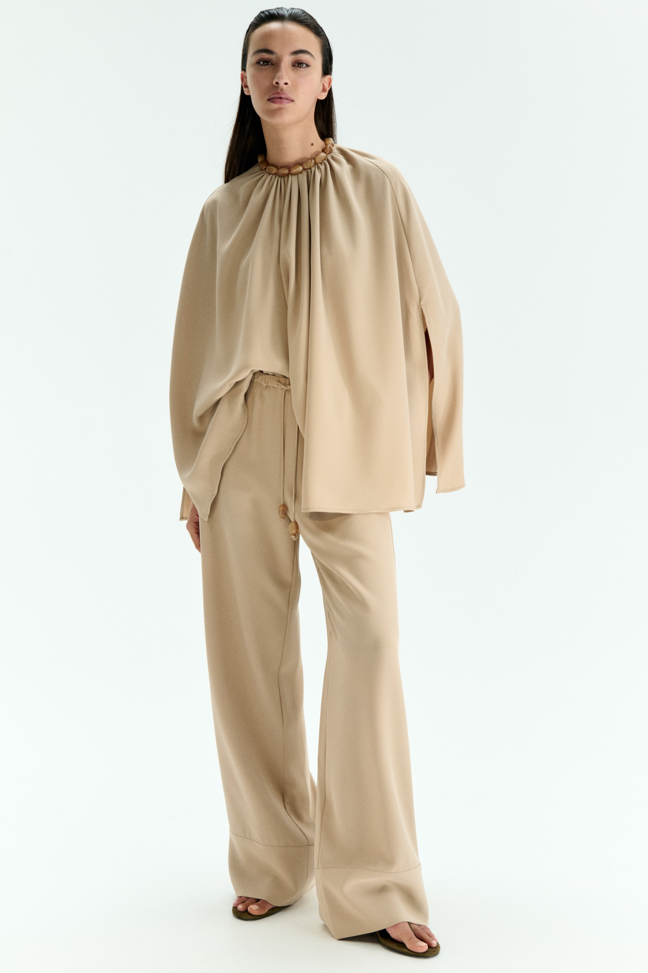 View larger image: A beige tunic and wide-leg pants set is being worn by the model. The tunic is featuring a gathered high neckline with wooden bead details and wide sleeves with side slits, while the wide-leg pants are featuring a drawstring waist with coordinating beaded ends.