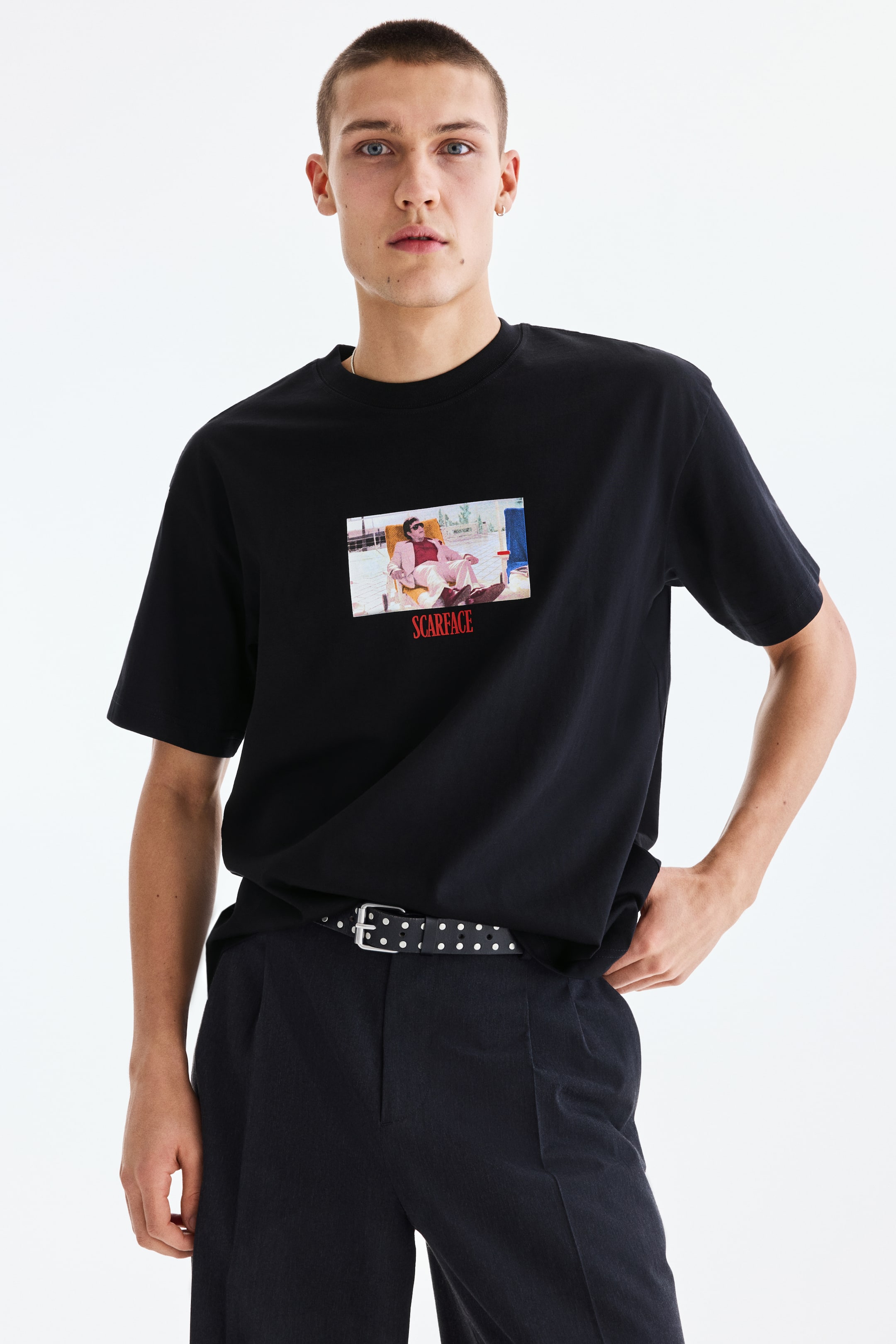 Ver imagem maior: Uma t-shirt preta apresenta uma gola redonda e mangas curtas vista de frente, exibindo uma estampa gráfica retangular centrada no peito. Esta imagem retrata uma pessoa num fato rosa e óculos de sol reclinada numa cadeira, com a palavra 'SCARFACE' em texto vermelho diretamente abaixo.