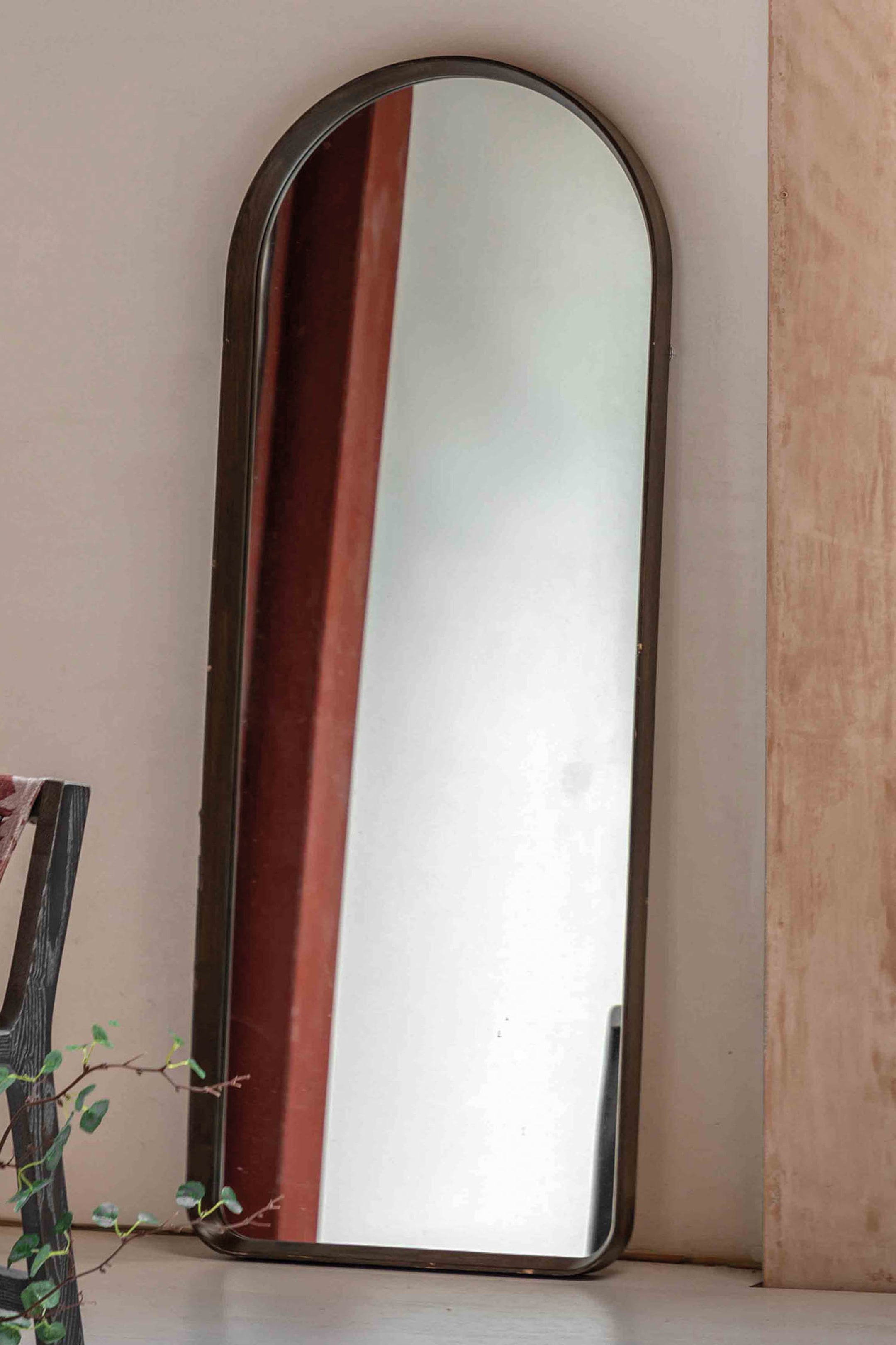 View larger image: A tall, arch-top mirror stands upright, angled slightly to its right, showcasing the depth of its slim, dark bronze-coloured metal frame.